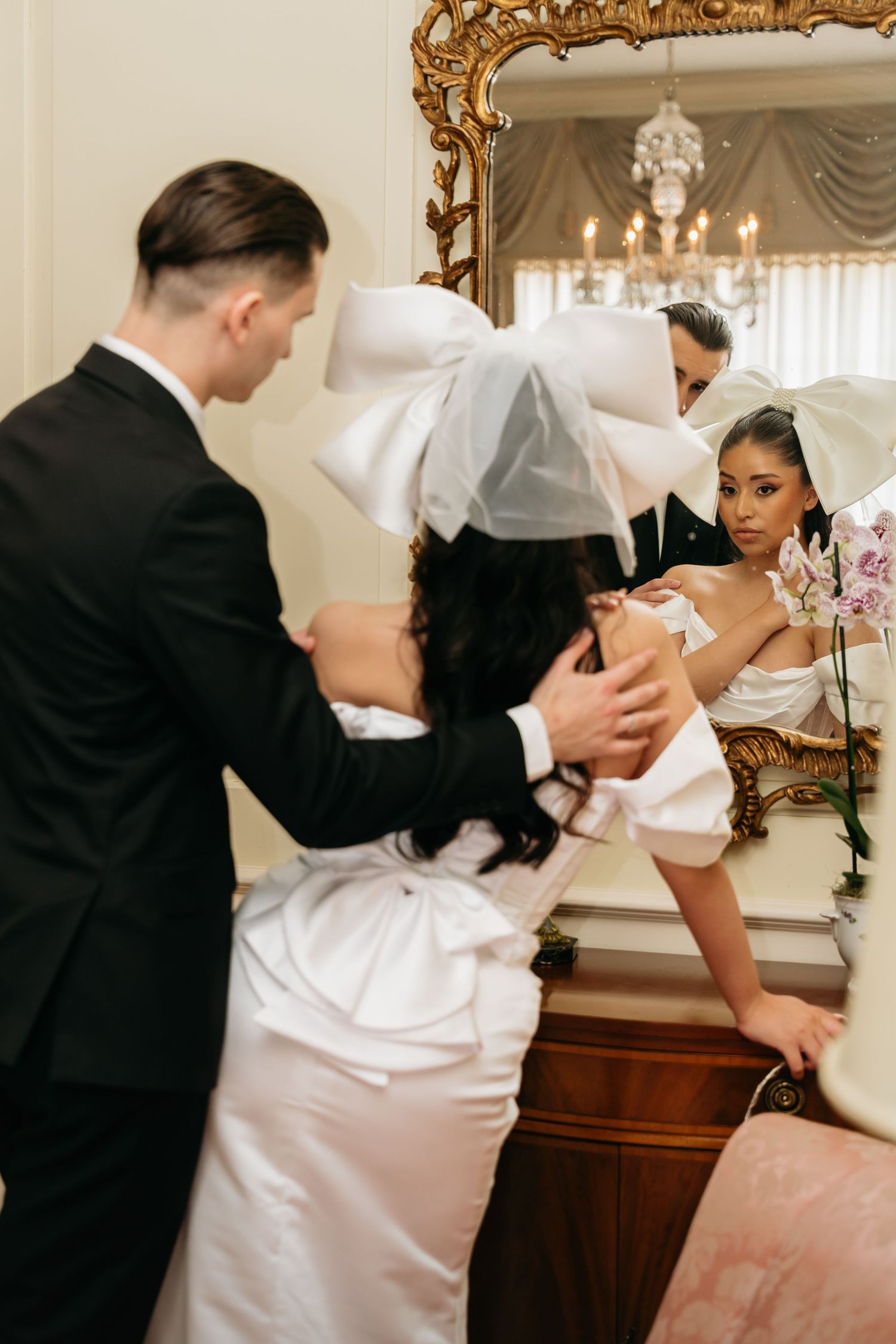 Groom in black tuxedo helping bride in white satin gown with large bow headpiece near ornate gold mirror.