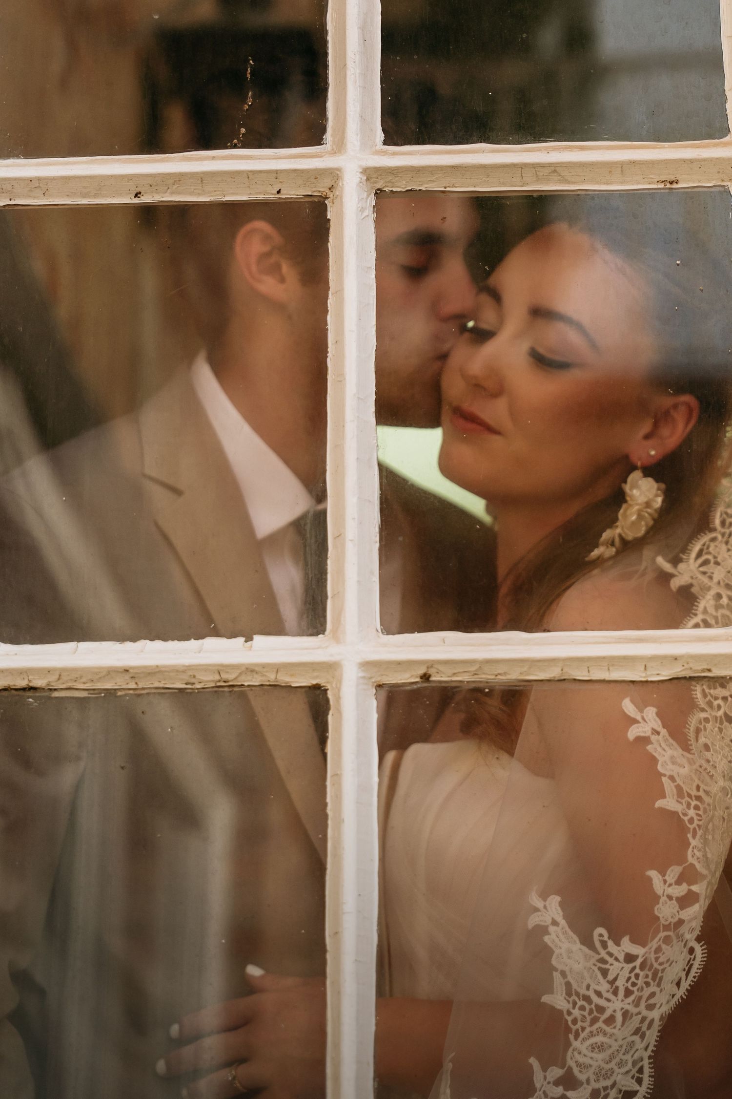A bride and groom share a romantic kiss through a vintage white-framed window, bride in lace gown with elegant earrings.