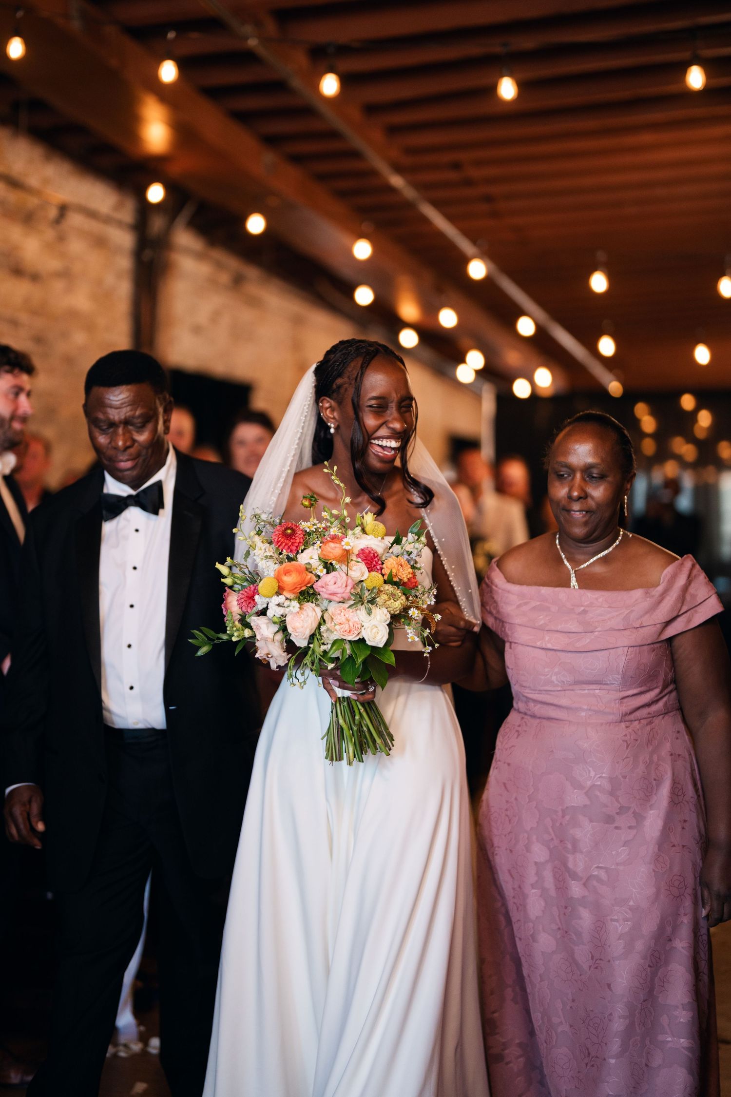 Radiant Black bride walks down aisle with parents under string lights holding colorful bouquet.