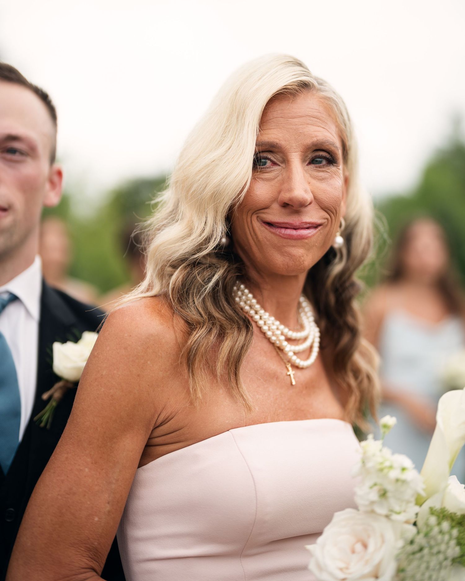 Emotional blonde woman in strapless blush dress and pearl necklace holding white flowers at a wedding ceremony.