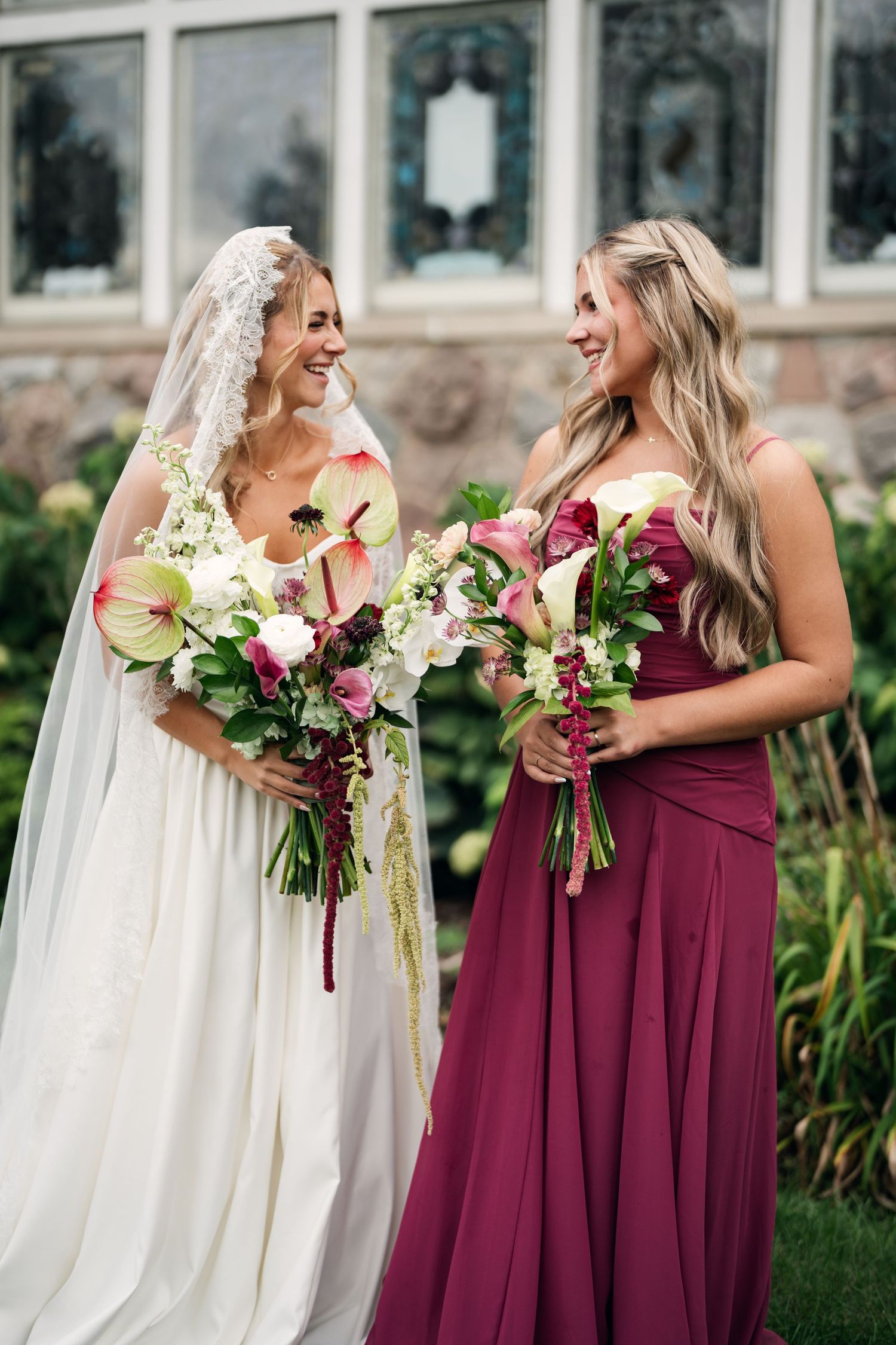 Bride in white gown and veil smiles with bridesmaid in burgundy dress, both holding floral bouquets outdoors.