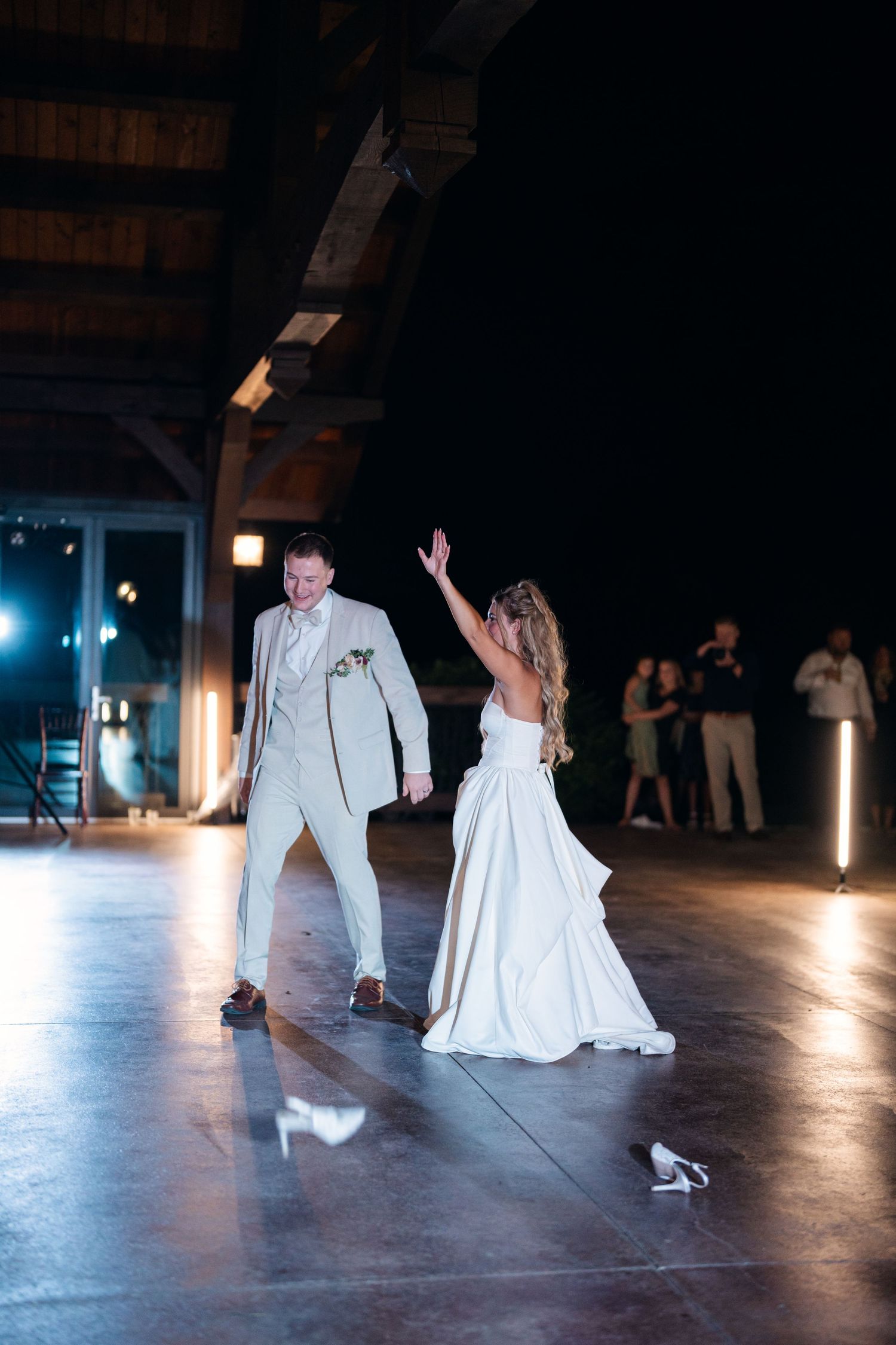 Bride and groom share their first dance in a dimly lit barn venue, bride raising her hand gracefully in a white gown.