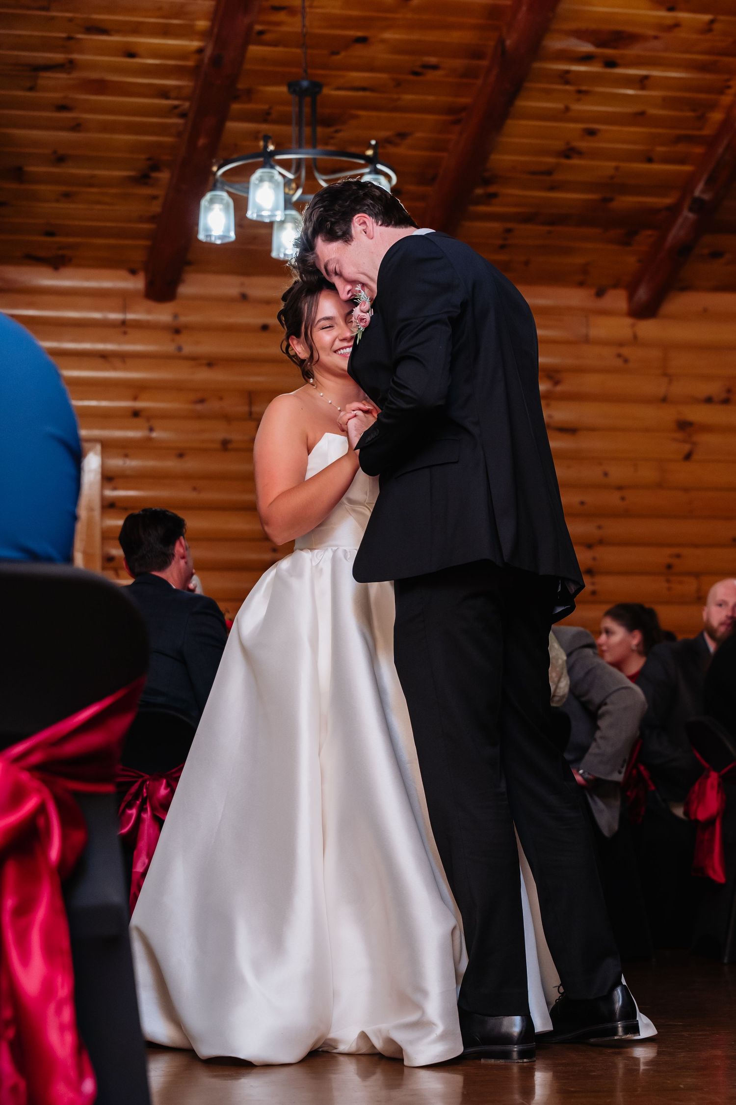 Bride and groom share their first dance in a rustic wood-paneled wedding venue with guests seated in background.