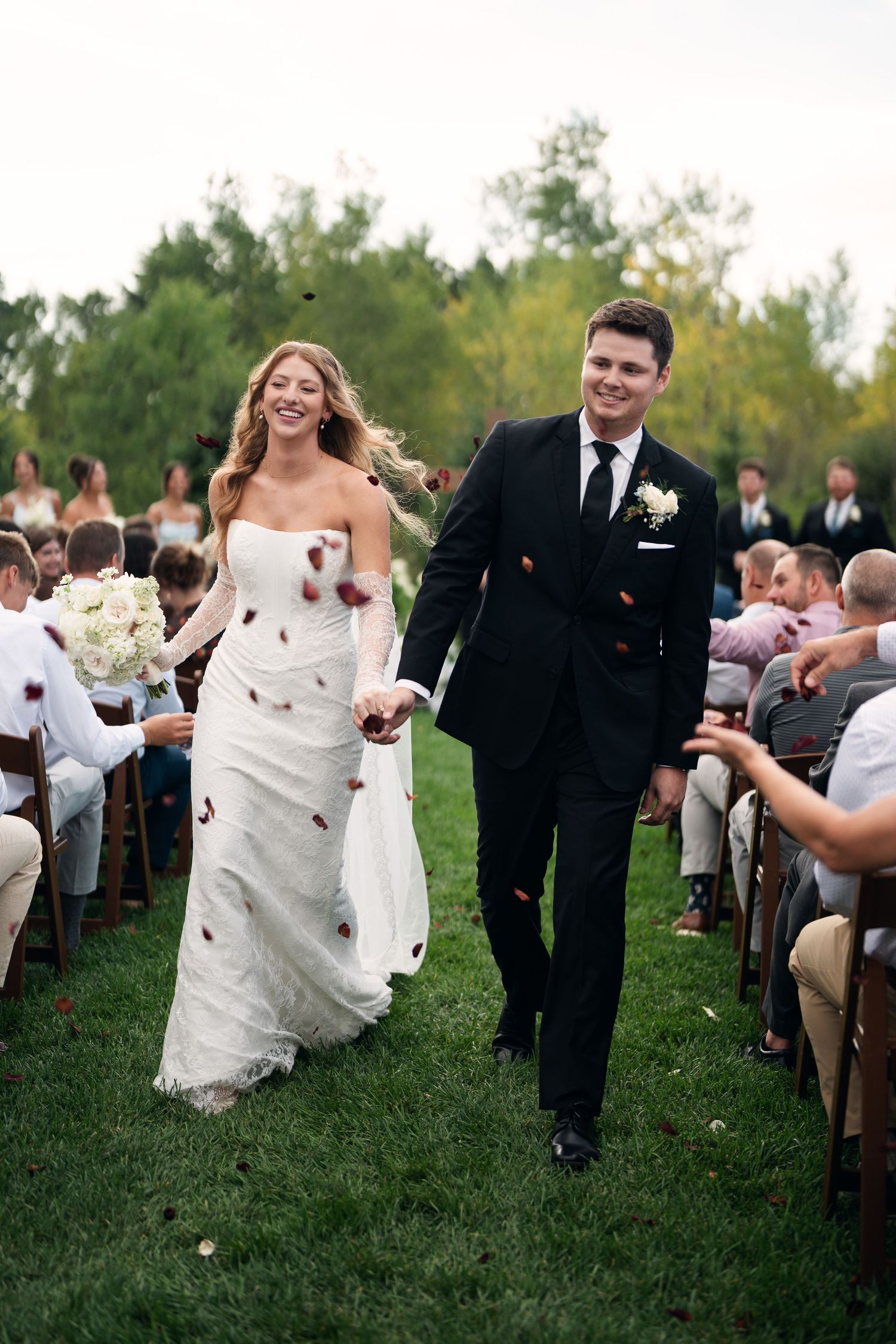 Joyful newlyweds walk down outdoor aisle as guests toss red rose petals in celebration.