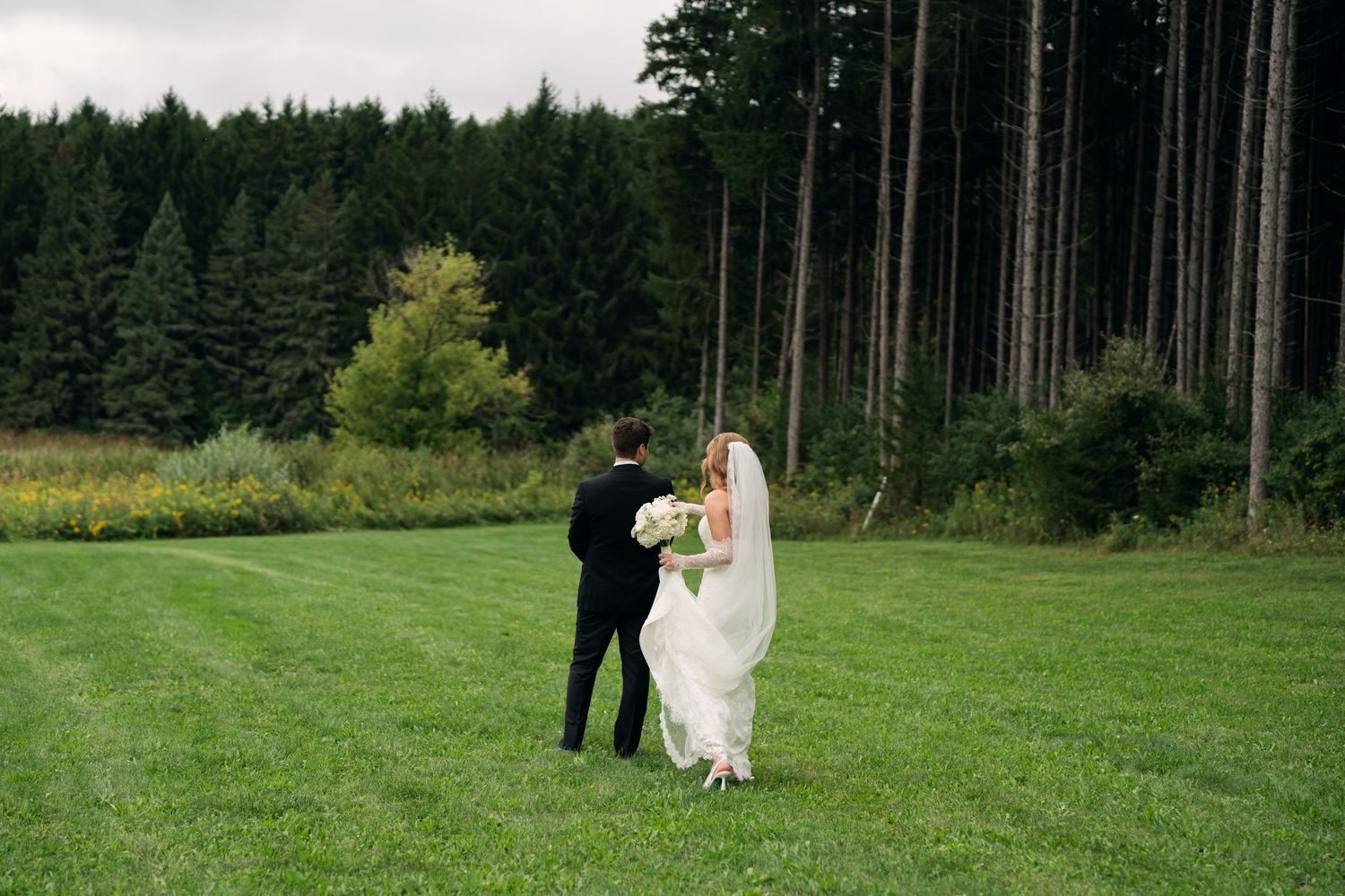 A bride and groom walk hand in hand across a lush green lawn with a dense evergreen forest backdrop.