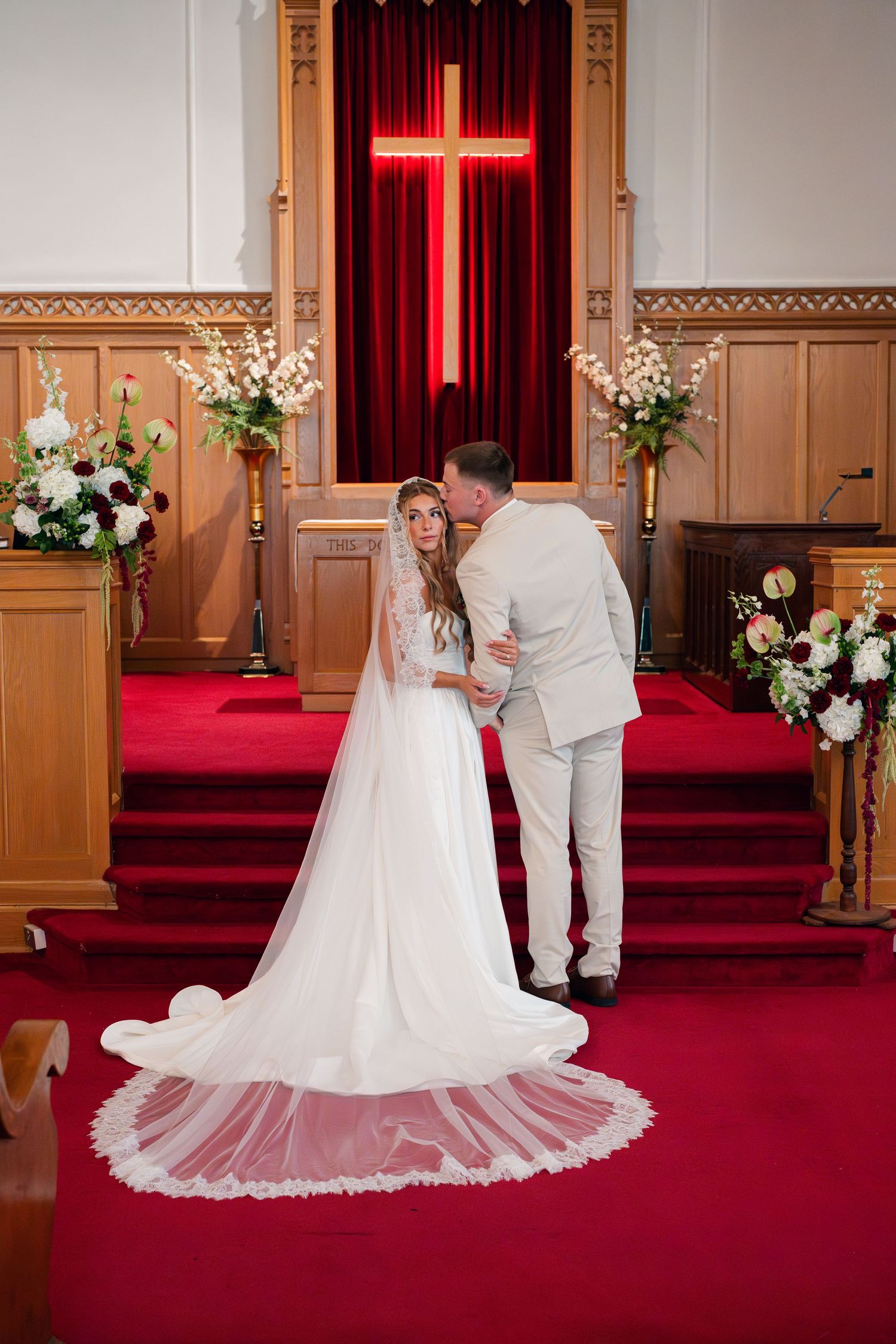 Bride and groom share a tender moment at the altar of a church decorated with floral arrangements and a glowing red cross.