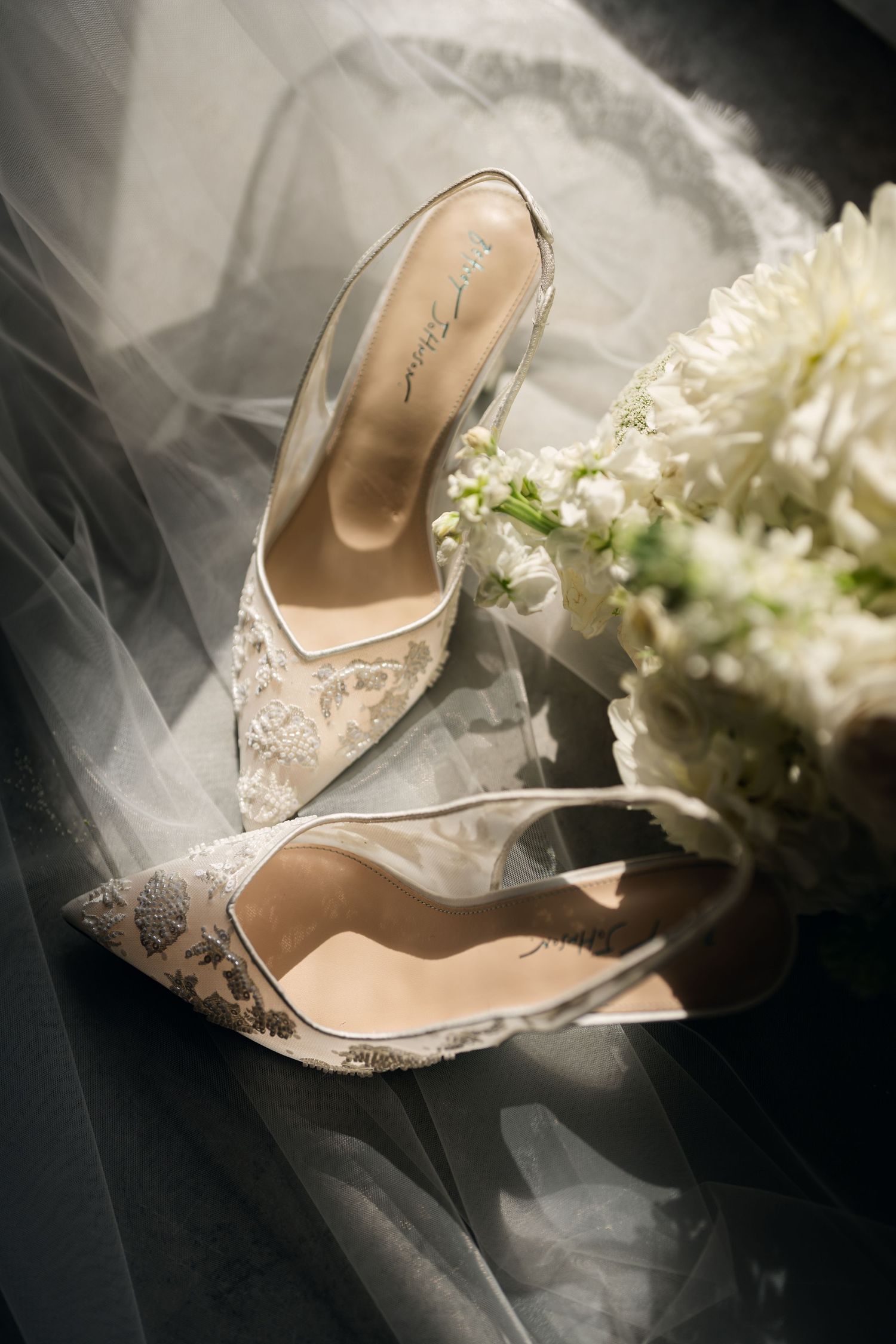 Elegant lace bridal heels with clear slingback straps resting on a white veil beside a bouquet of white flowers.