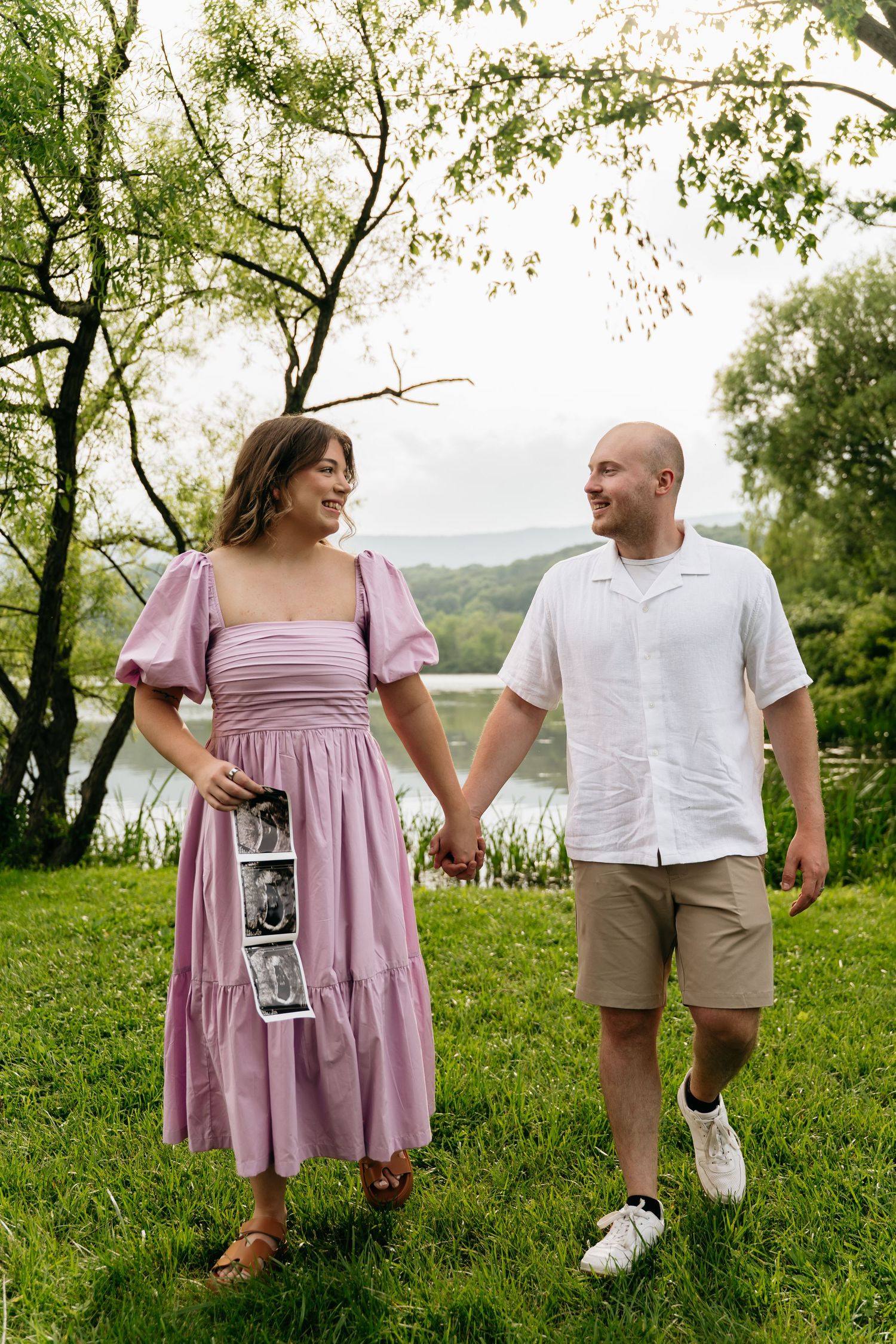 A couple stands together on a grassy hillside, with her in a pink dress and him in a white shirt and khaki shorts.