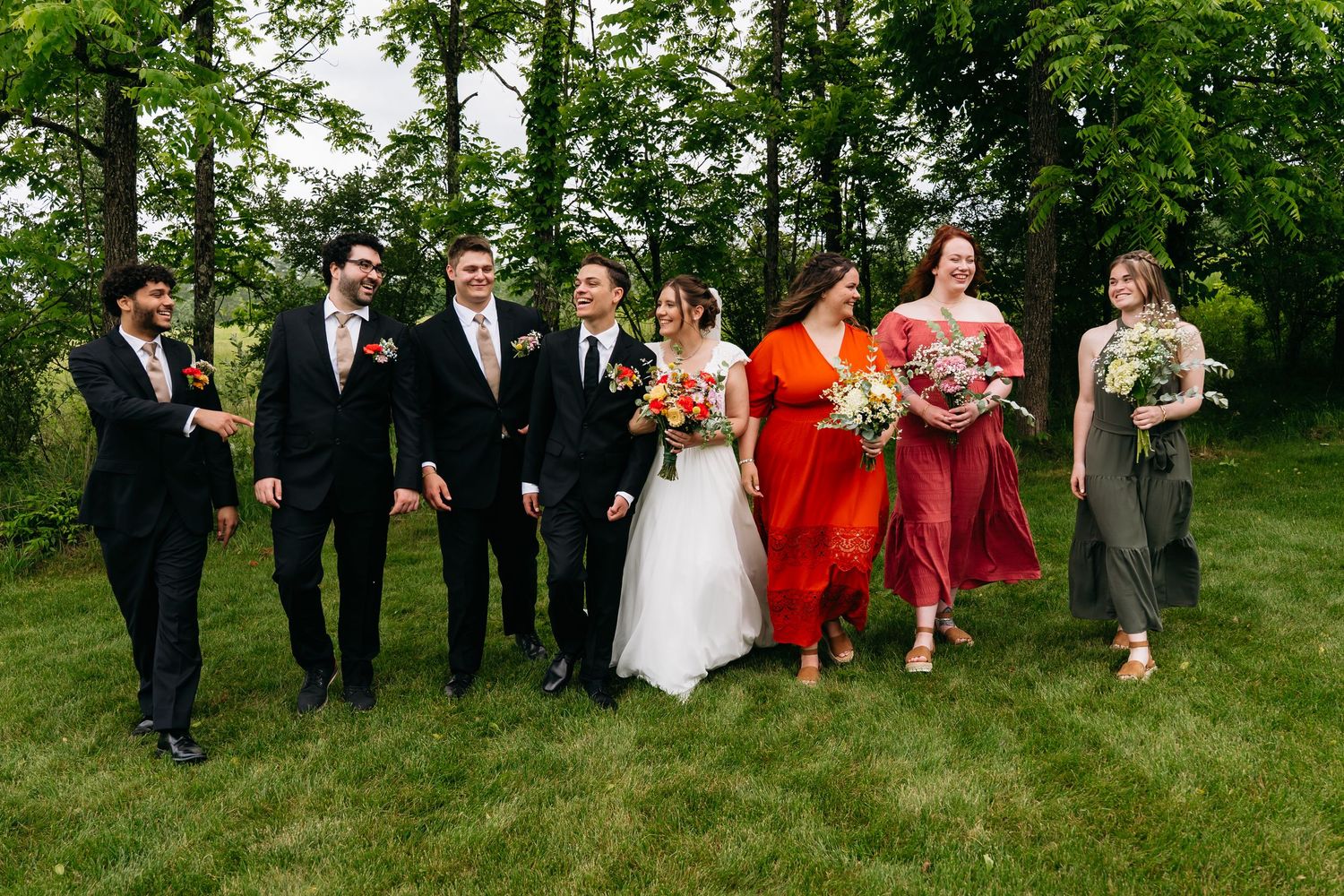 A wedding party wearing red bridesmaid dresses and dark suits poses together on a grassy lawn in natural light.