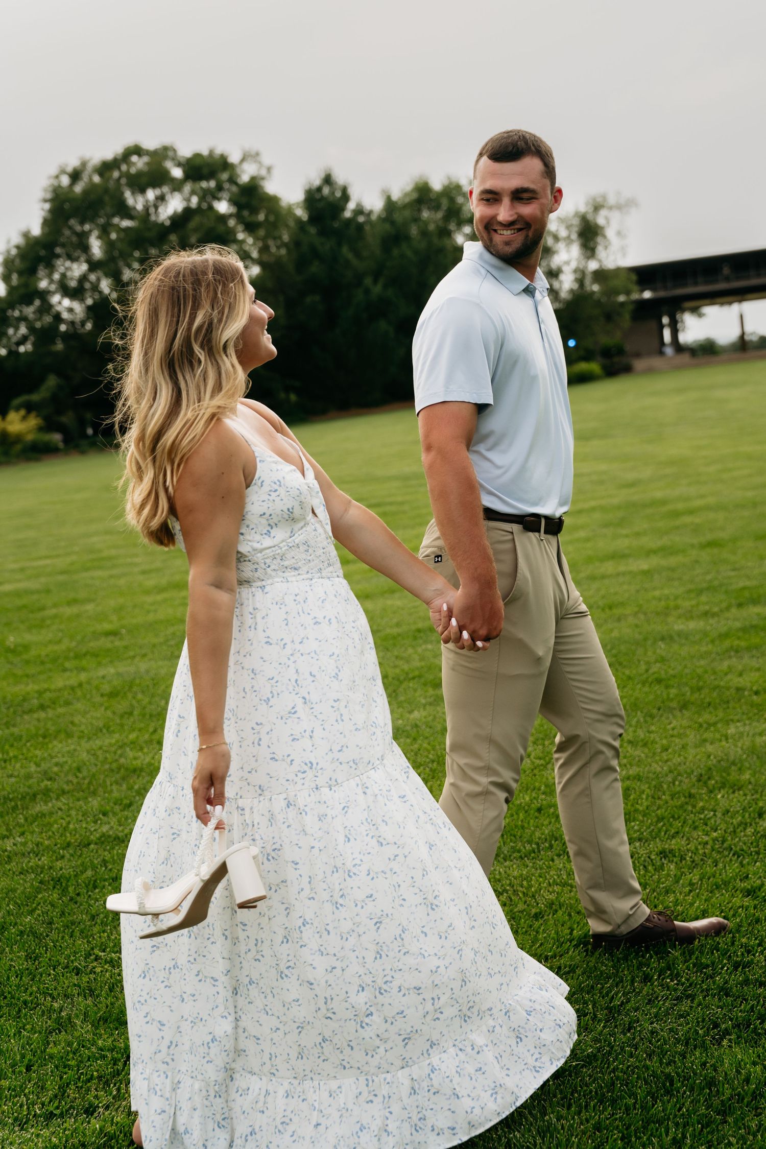 A couple walks hand in hand across a lush green lawn, with the bride wearing a flowing white floral dress.
