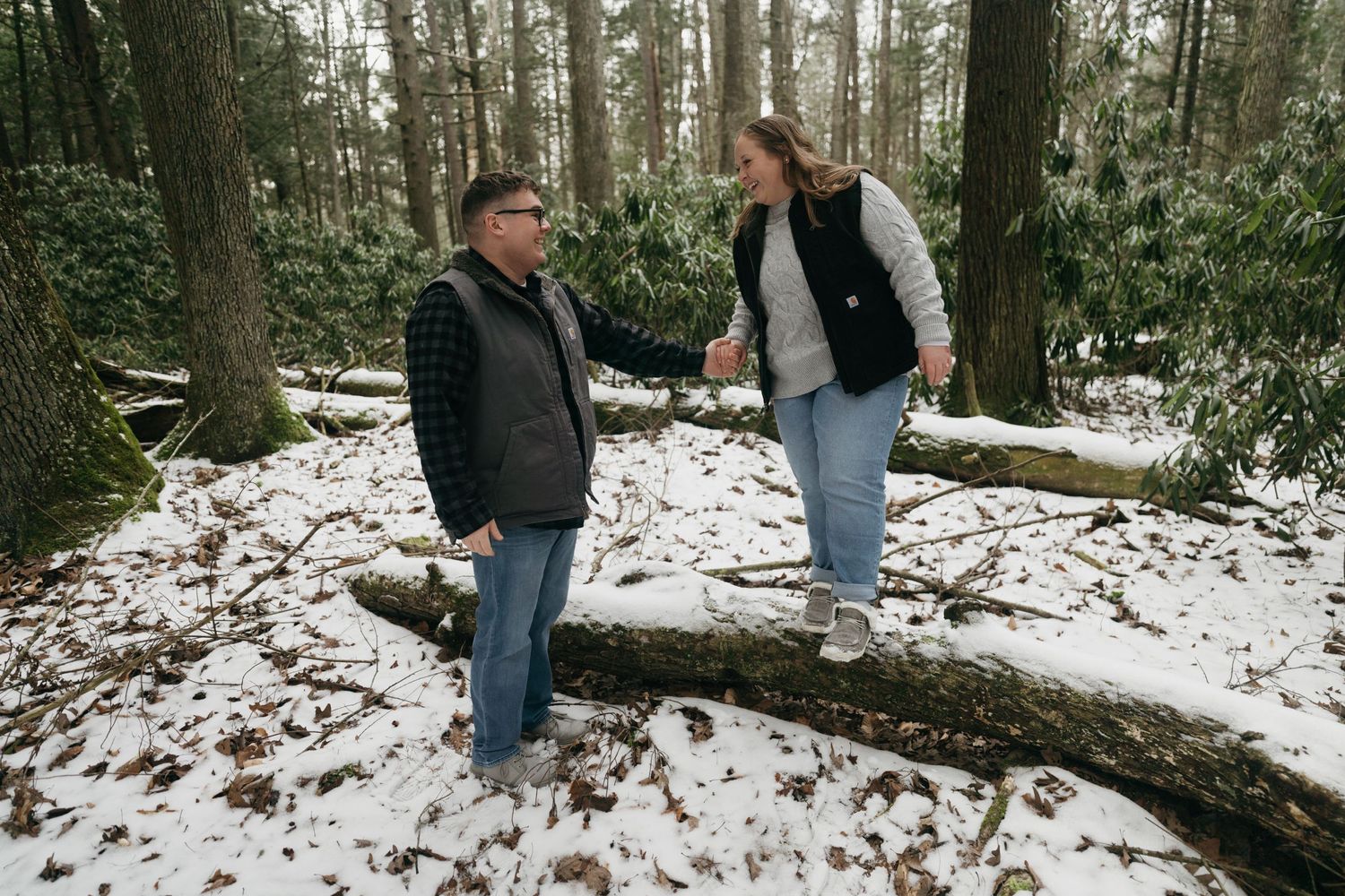 A couple having fun while standing on a fallen tree in snowy woods.