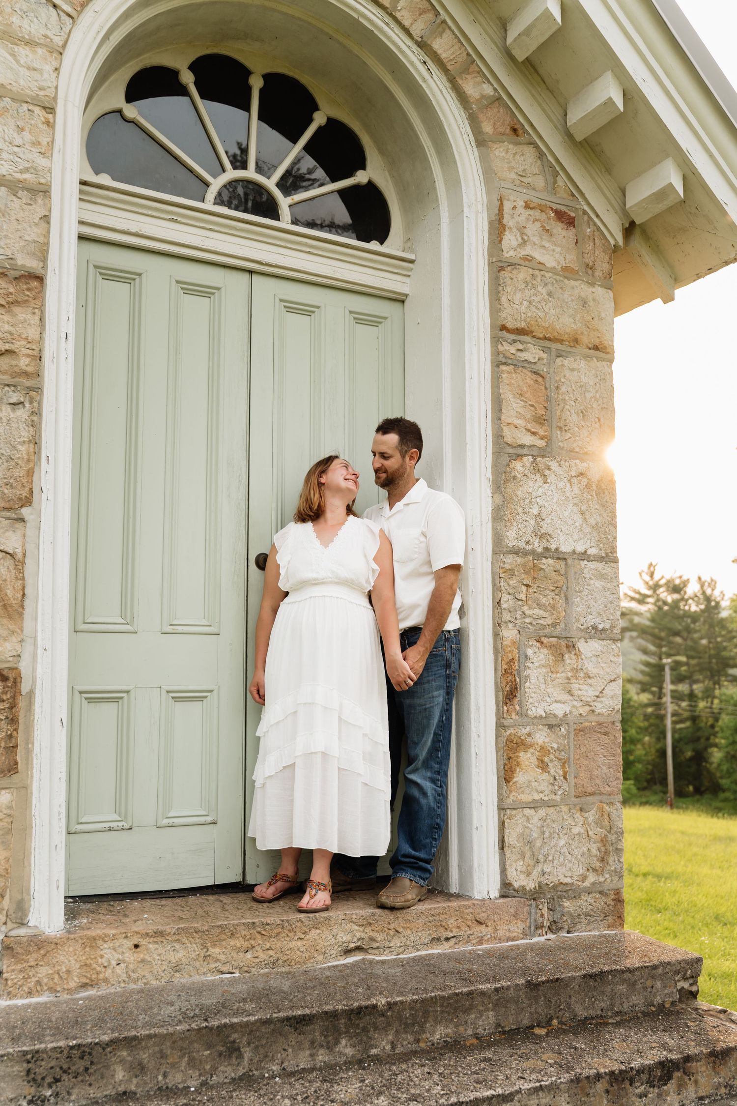 A couple stands intimately in the doorway of a historic stone church with an arched entrance at sunset.