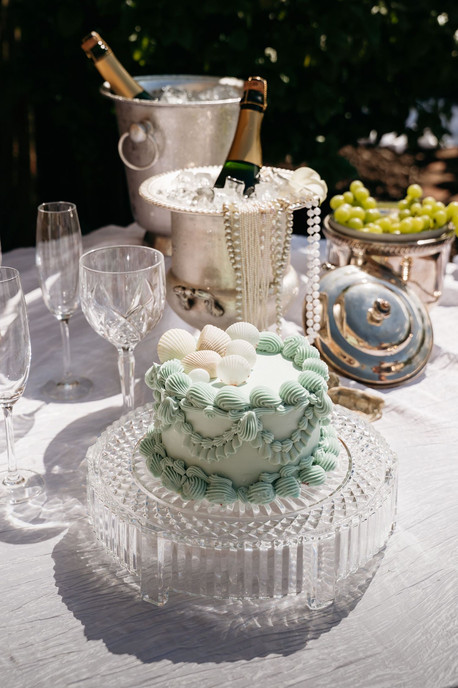 Elegant wedding cake with mint green frosting on crystal stand beside champagne bottles and glasses.