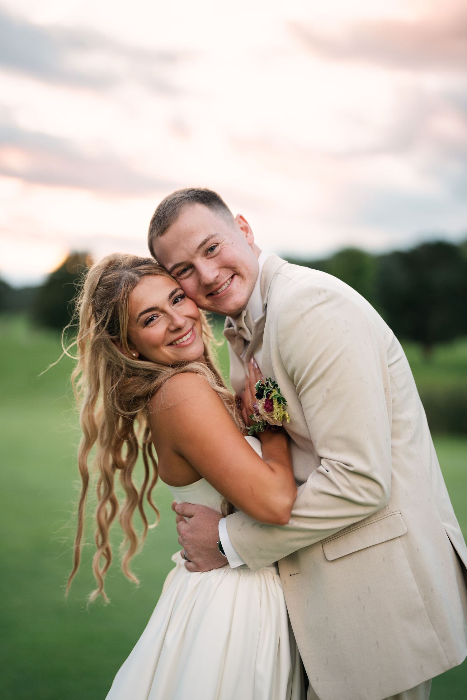 Smiling bride and groom embrace warmly on a golf course at sunset, both radiating joy and happiness.
