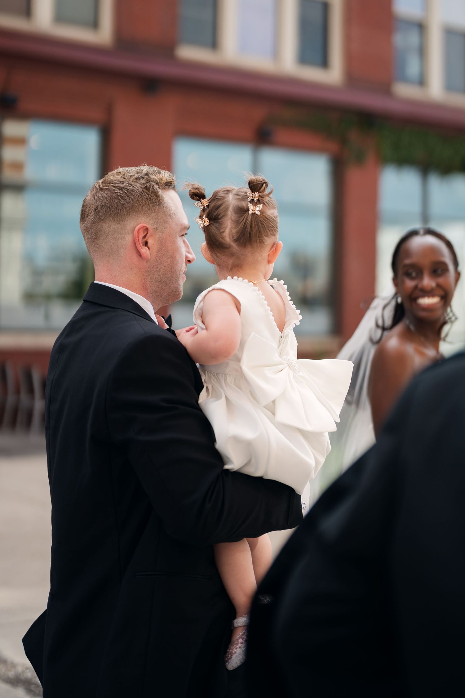 Groom in black suit holds young flower girl in white dress while smiling bride stands nearby at wedding.