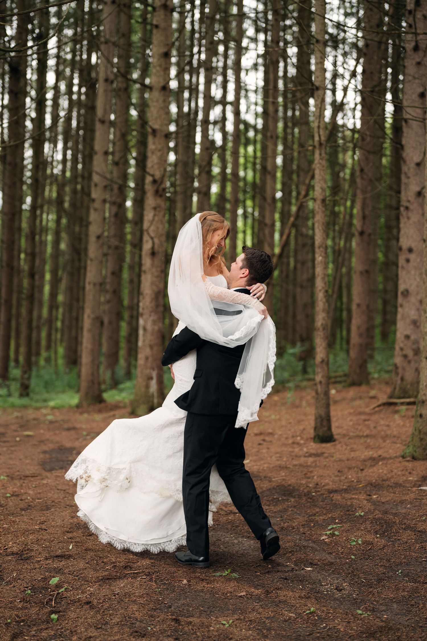 Groom lifting bride in white gown and veil sharing a romantic kiss in a lush pine forest.