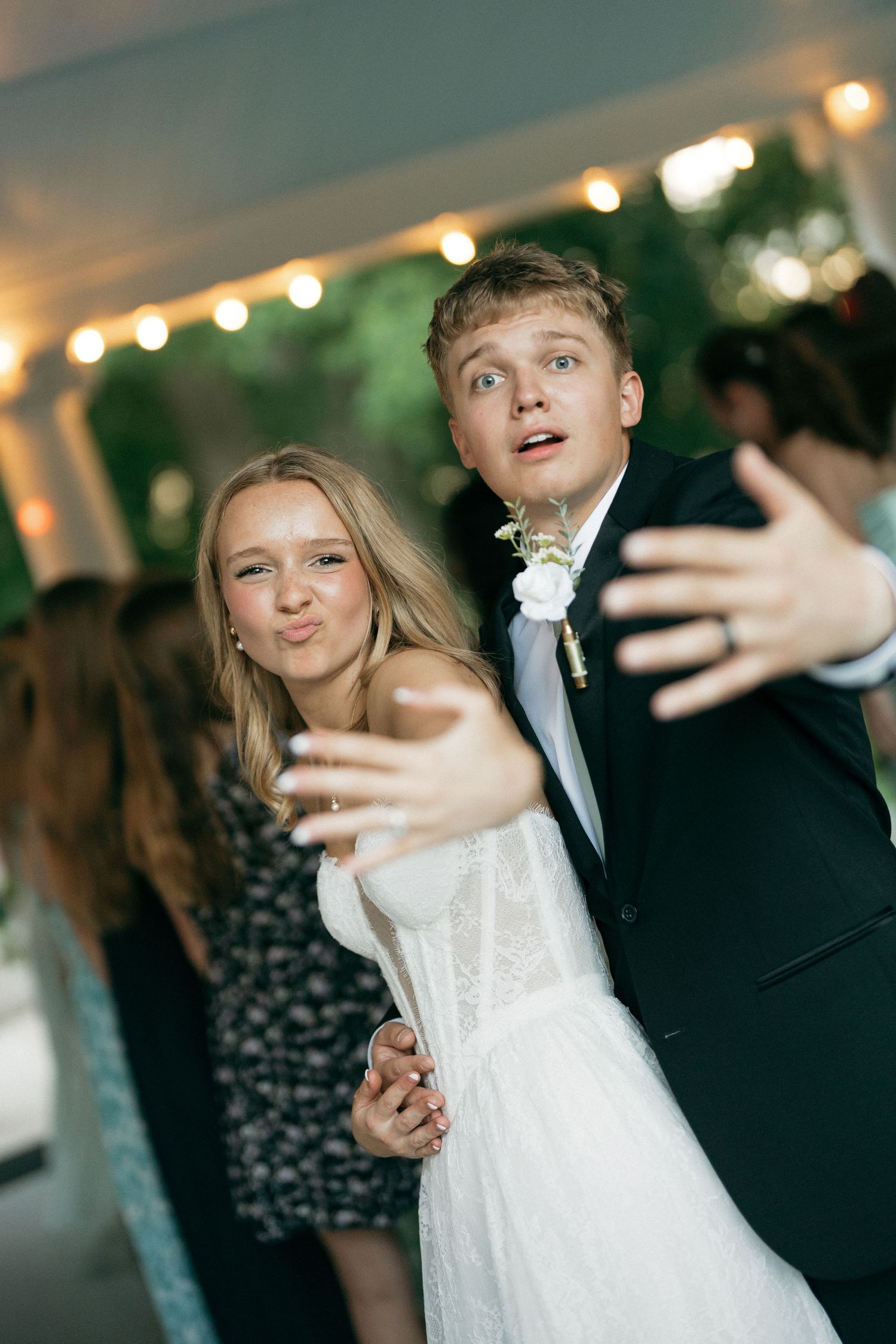 Young couple in formal attire dancing and posing playfully at an outdoor wedding reception under string lights.