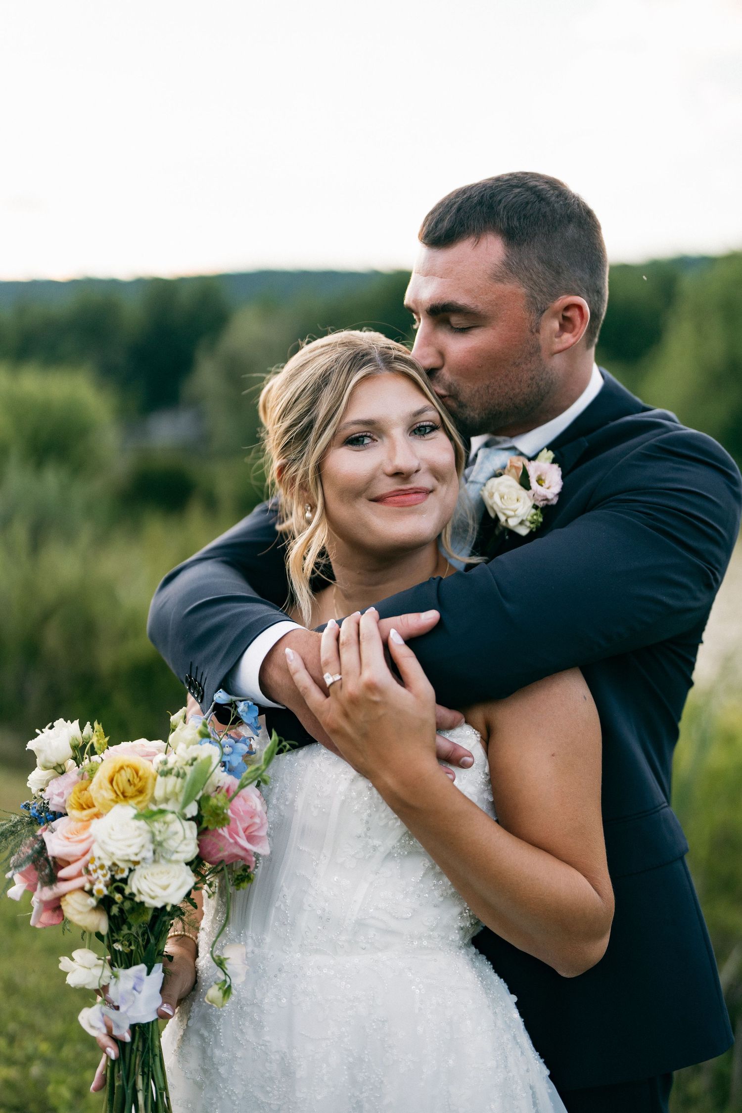 Groom in navy suit embracing bride in white dress while kissing her cheek at golden hour.