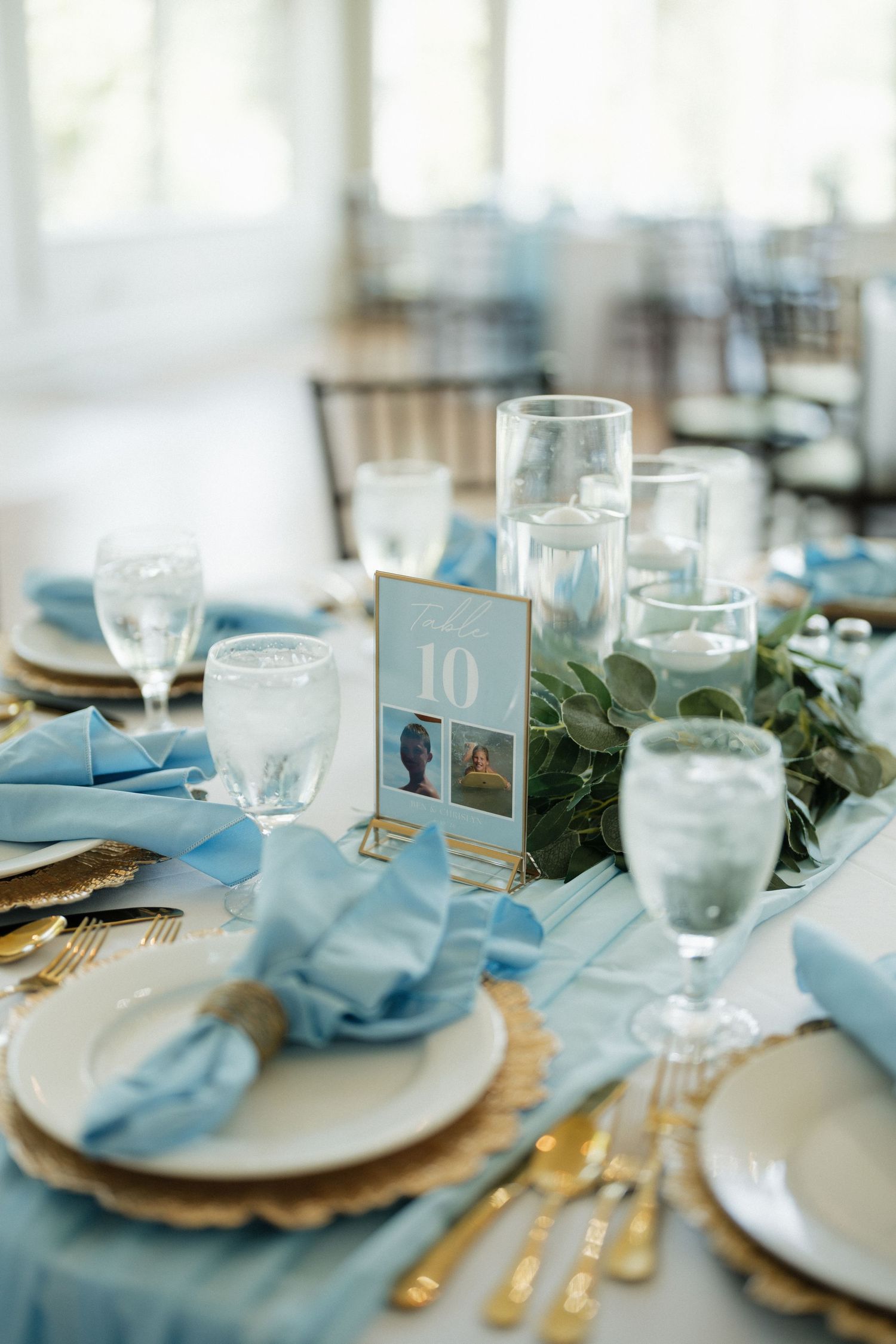 Elegant wedding reception table with blue napkins, gold chargers, floating candles, greenery centerpiece, and table number 10 card.