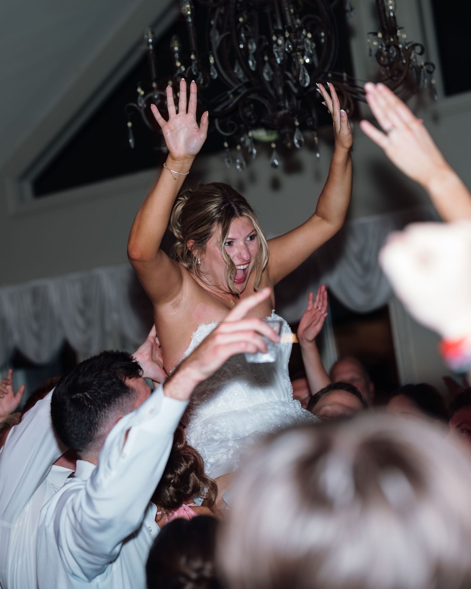 Excited bride being lifted by guests during lively wedding reception celebration indoors.