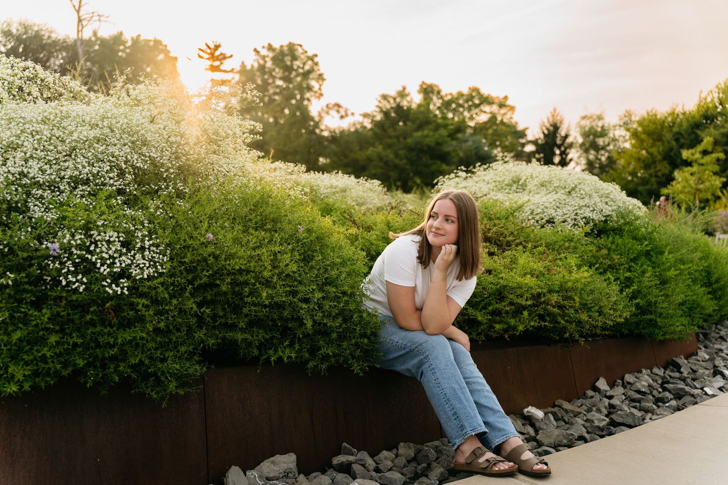 Young woman in white tee and jeans sits among white flowering shrubs at golden hour in a lush garden.
