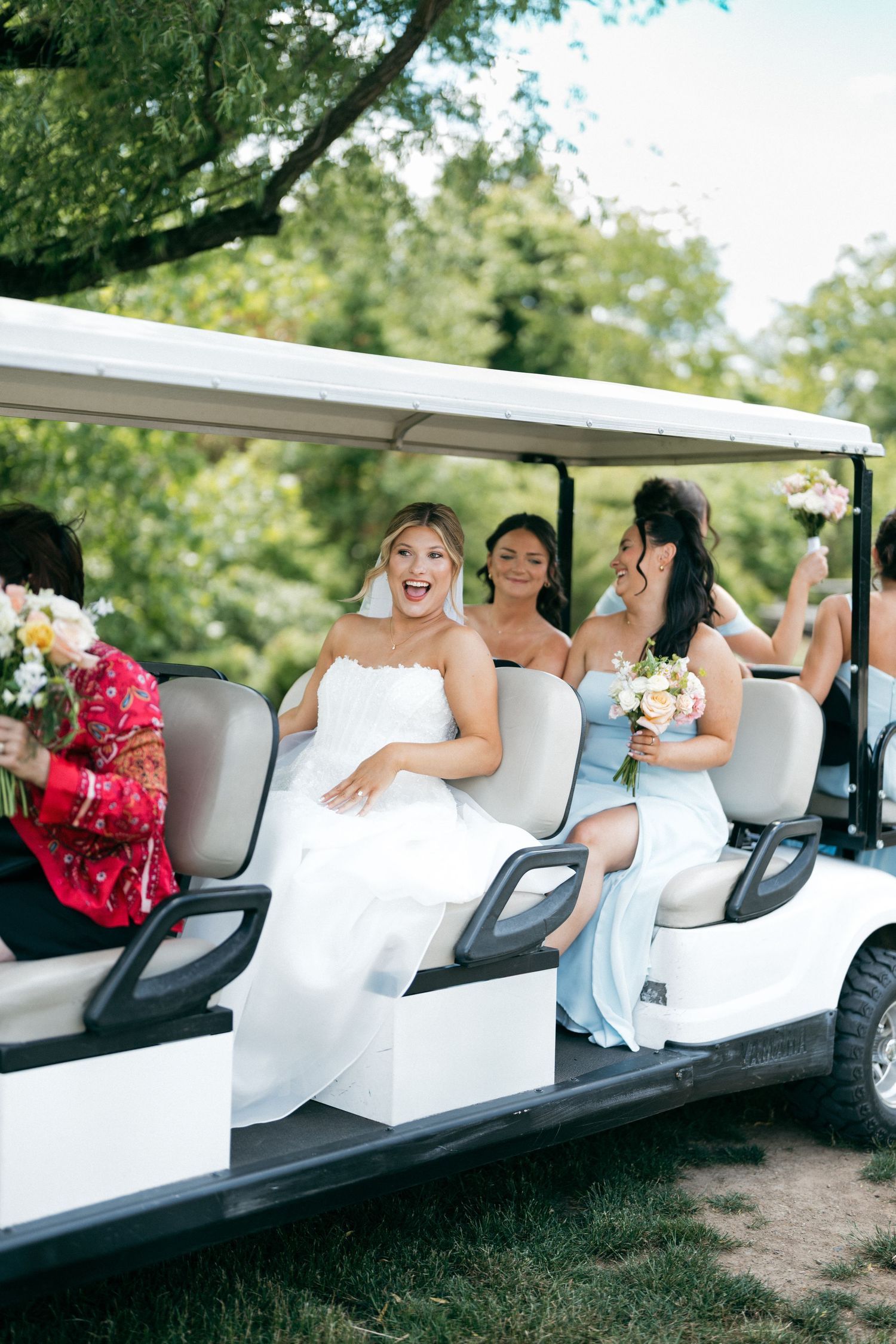 Bride and bridesmaids riding a golf cart outdoors, laughing and holding colorful bouquets.