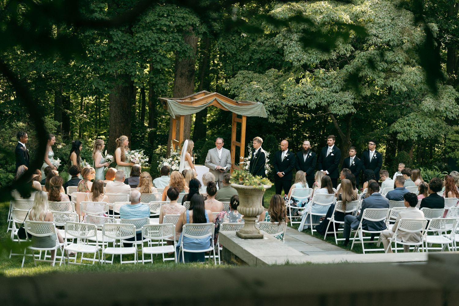 Outdoor wedding ceremony in lush green forest setting with white chairs and wooden arch altar.