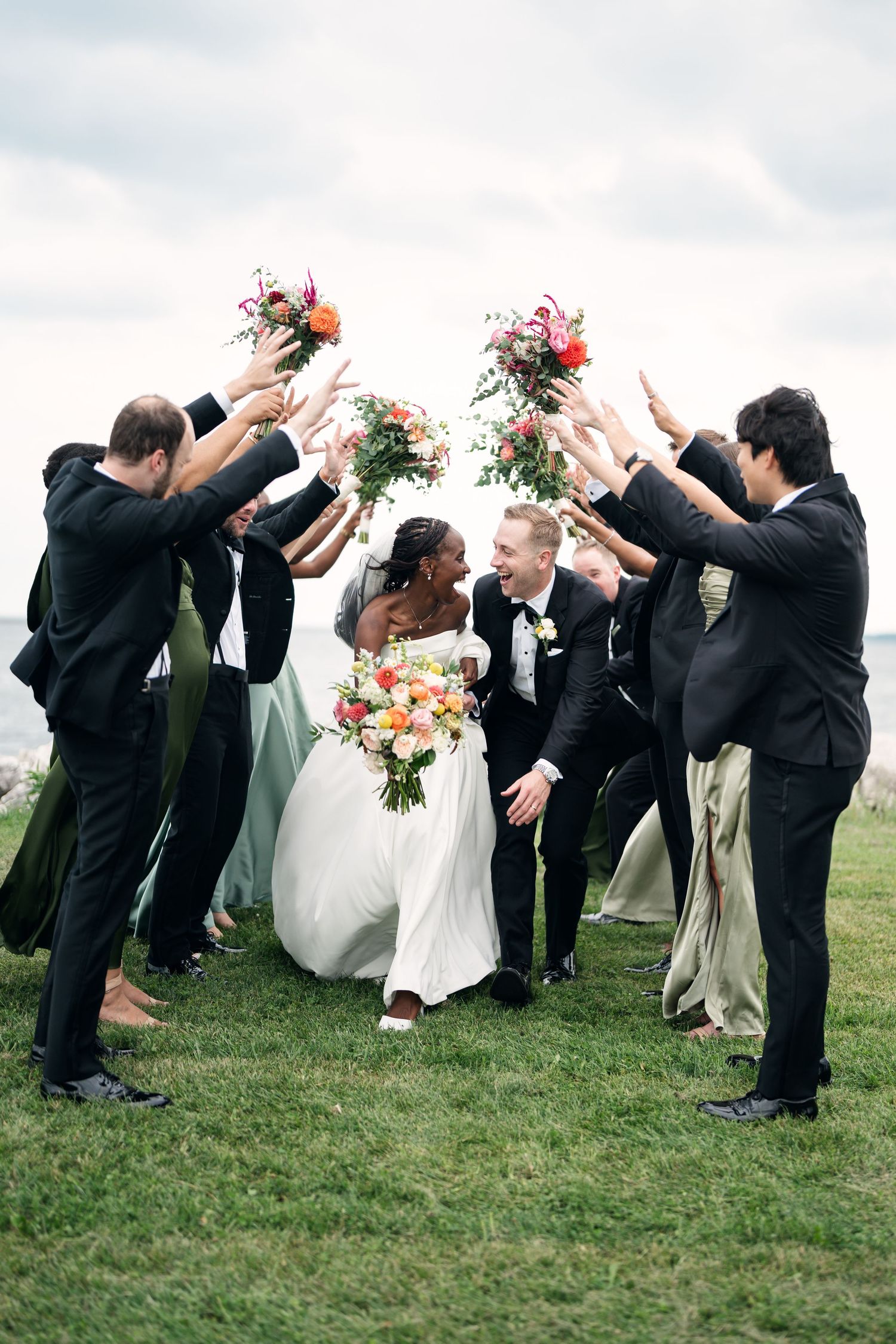Joyful interracial bride and groom walk through arch of raised bouquets held by wedding party on green lawn.