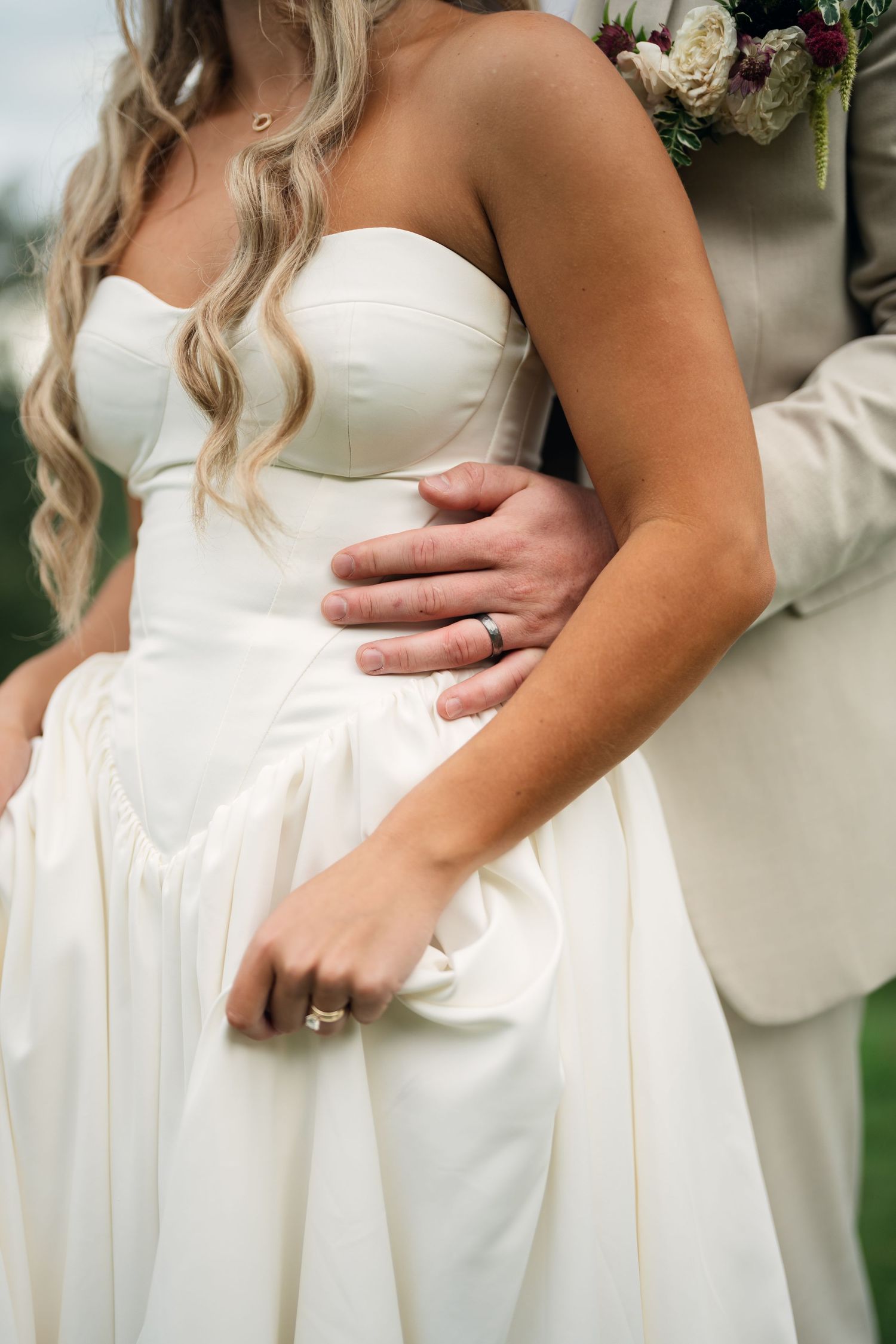 Bride in strapless white gown with groom's hand on her waist, both wearing wedding rings, holding a floral bouquet.