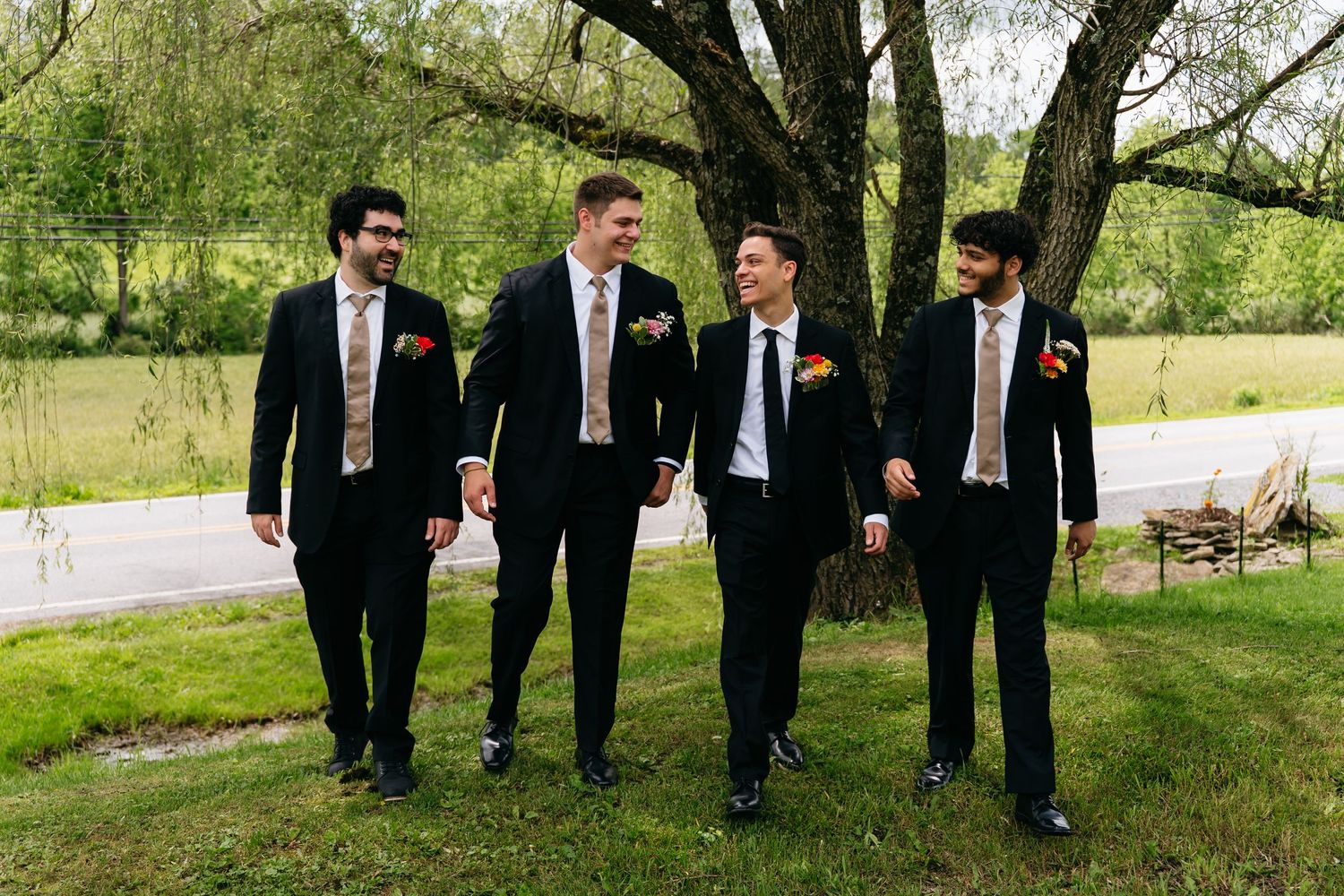 A group of four groomsmen in black suits walk and laugh together on a lush green lawn at an outdoor wedding venue.