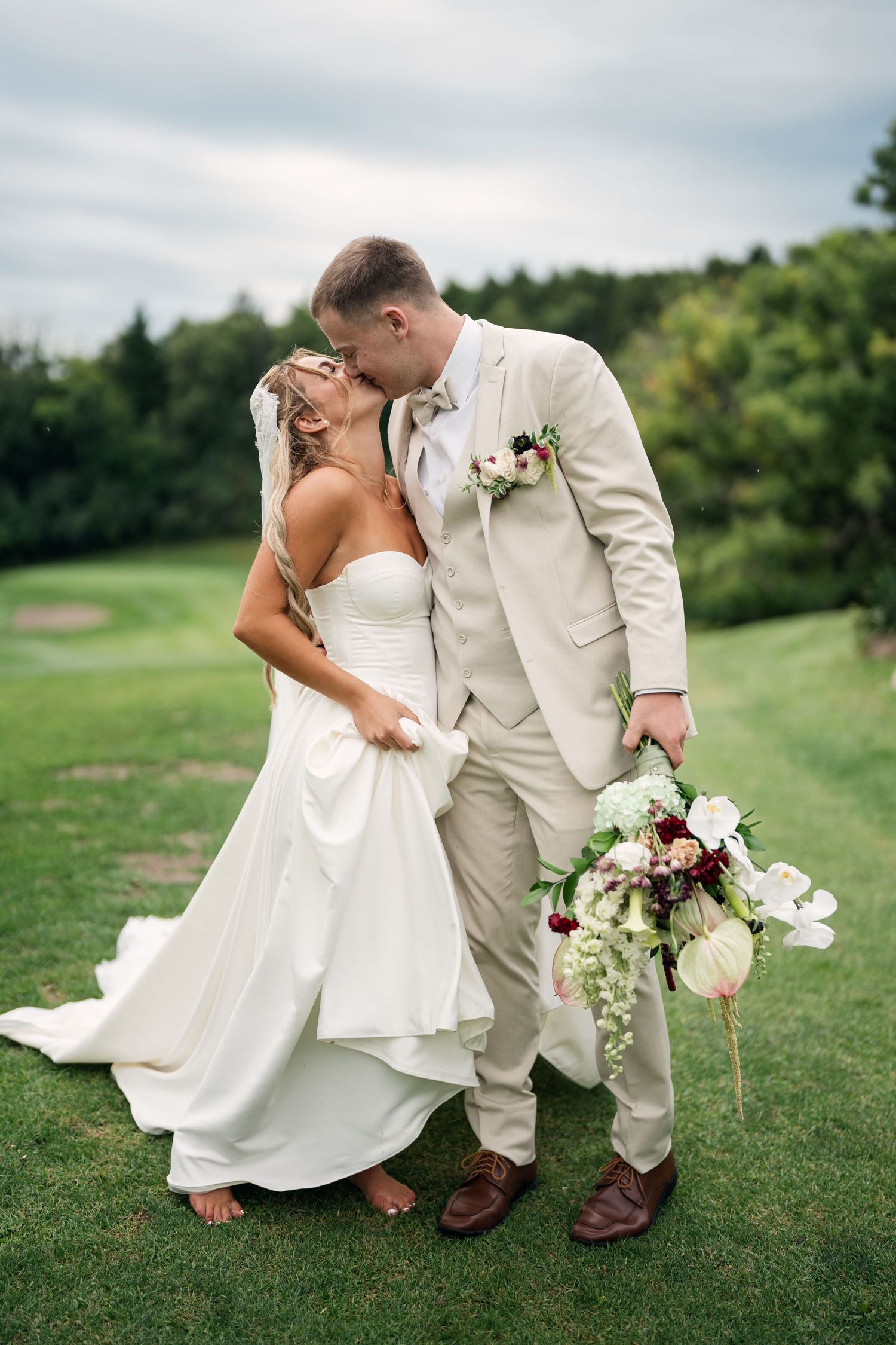 Bride and groom share a romantic kiss on green lawn, groom holds floral bouquet in beige suit.