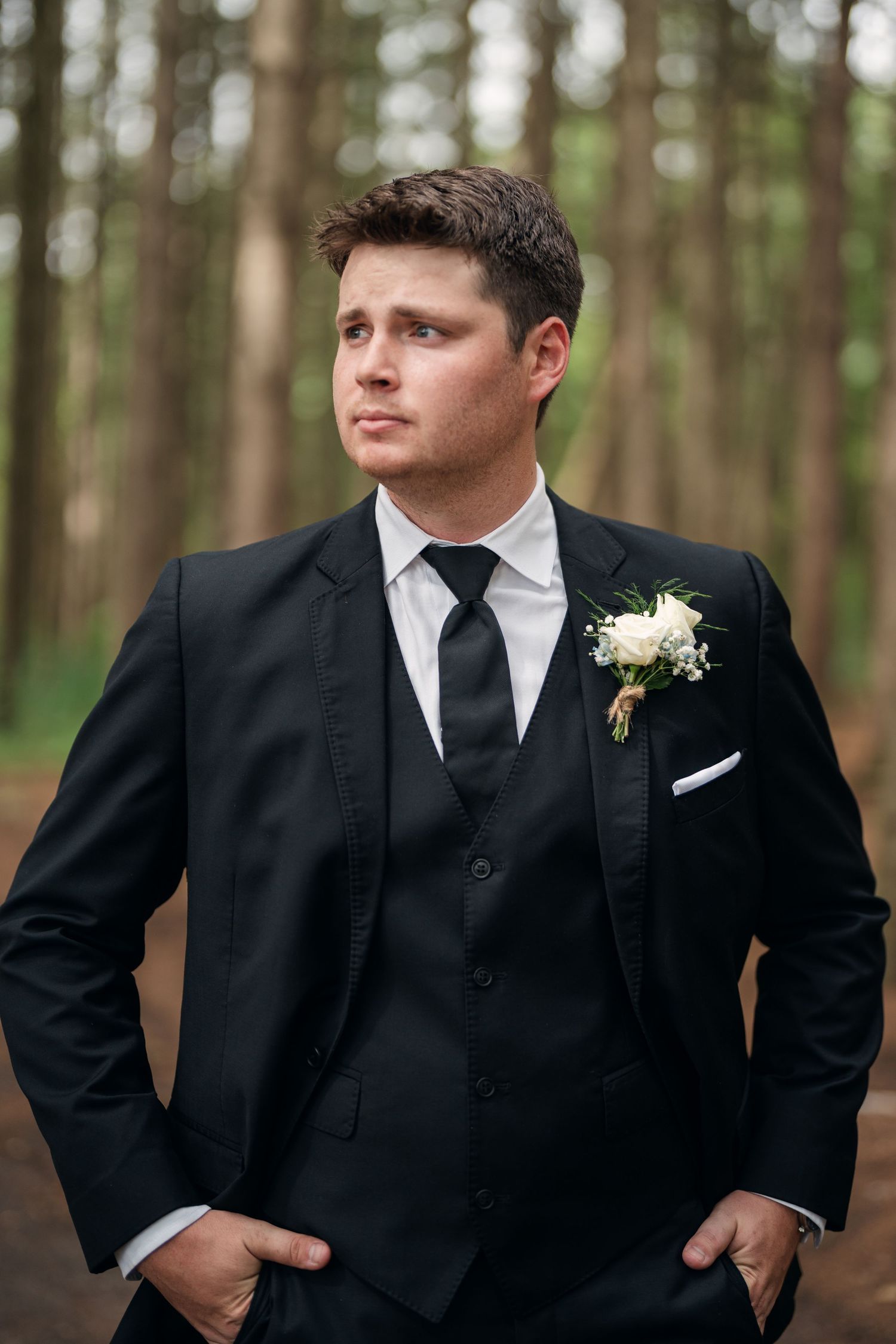 Groom in black three-piece suit with floral boutonniere standing in pine forest.