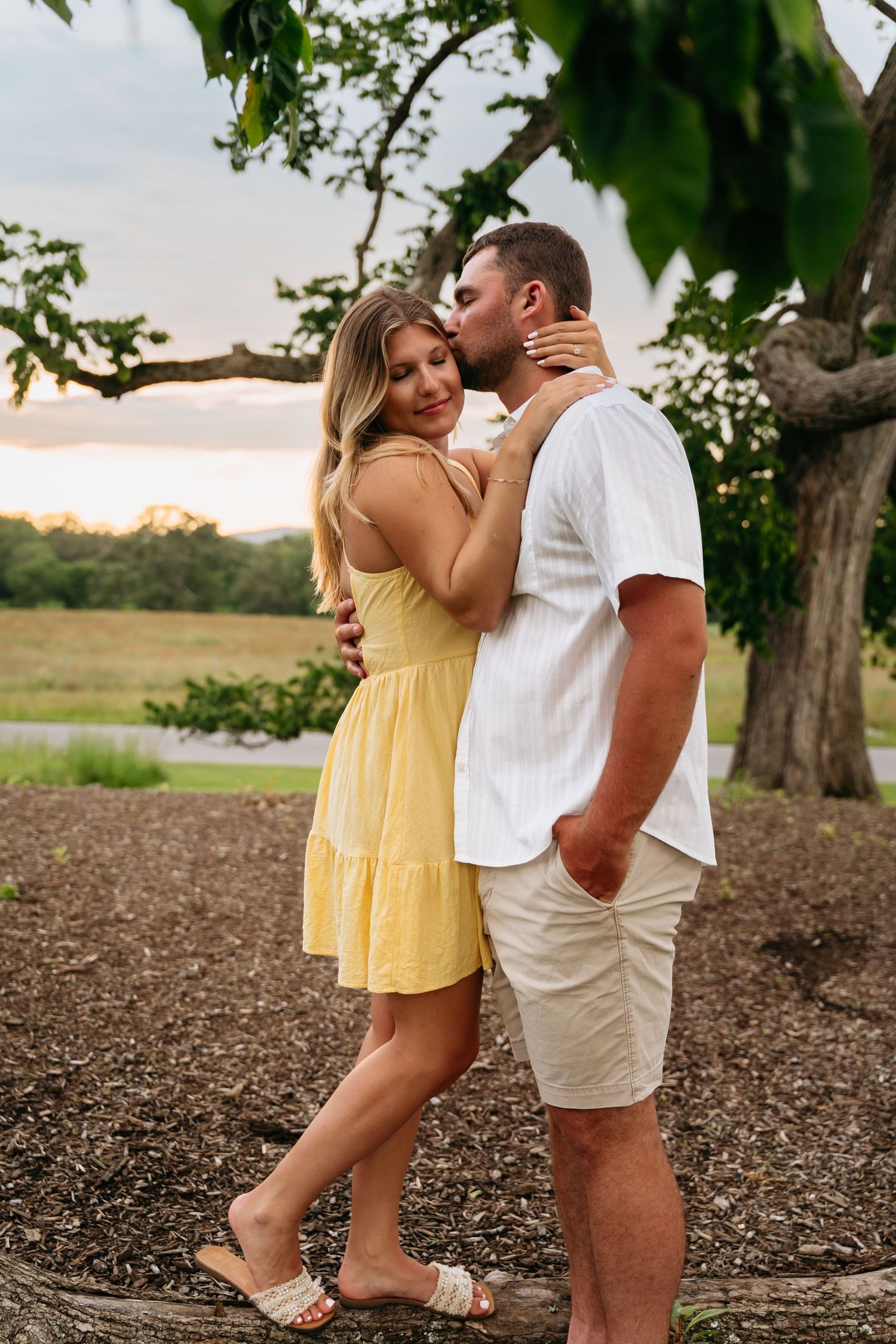 A couple shares a romantic moment under a tree at sunset in a rural setting, wearing yellow and white outfits.