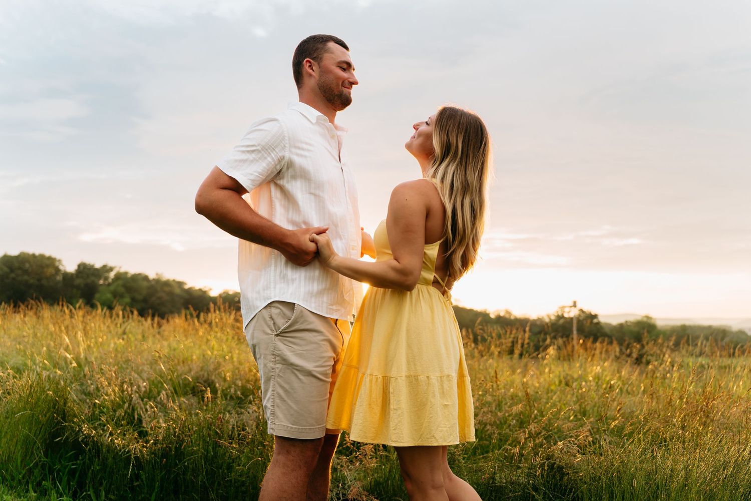 A couple holds hands while walking through a golden meadow at sunset in romantic countryside setting.