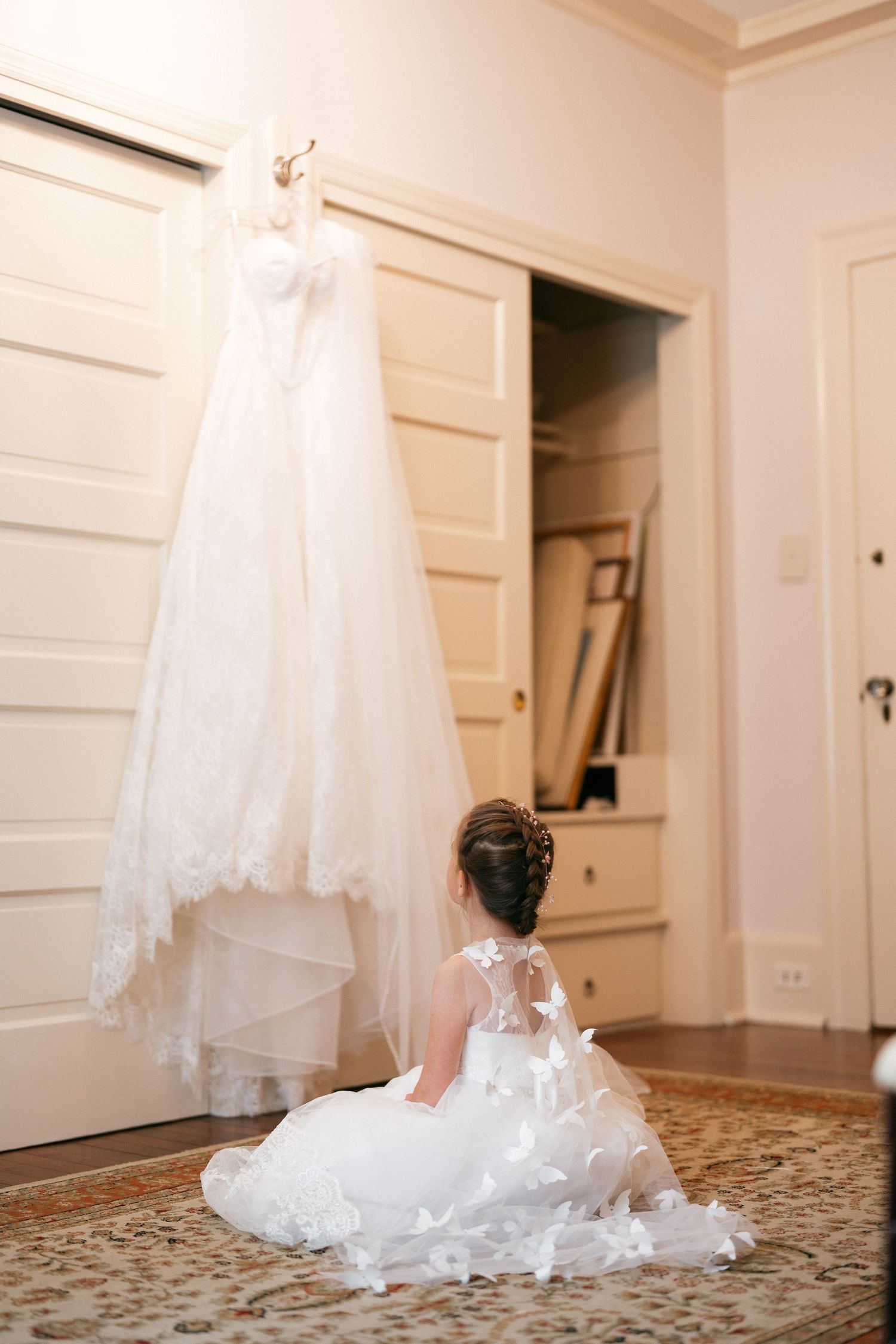 Flower girl in white dress sits on floor gazing up at elegant bridal gown hanging in doorway.