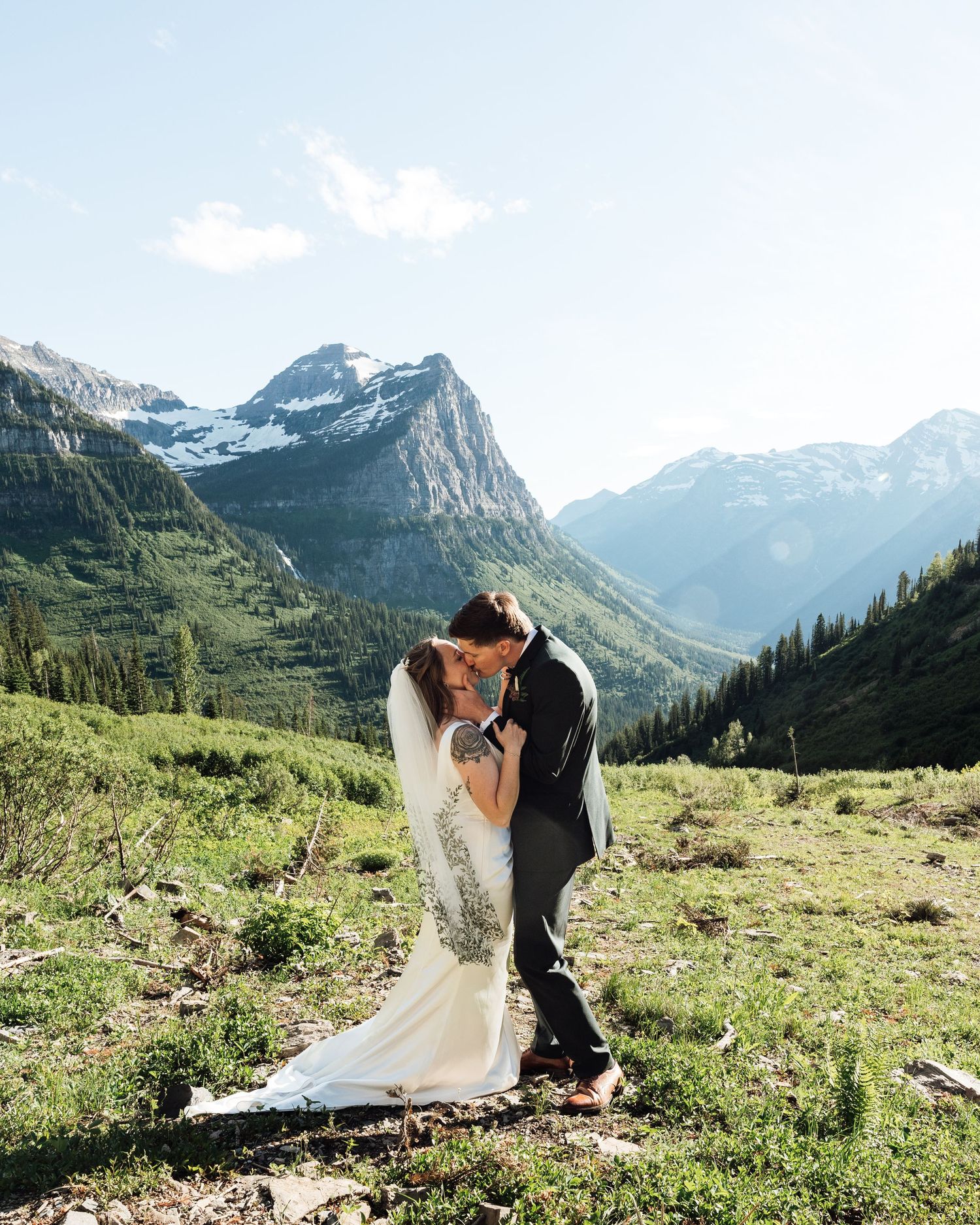 Bride and groom share a romantic kiss in a stunning mountain valley with snow-capped peaks and lush greenery.