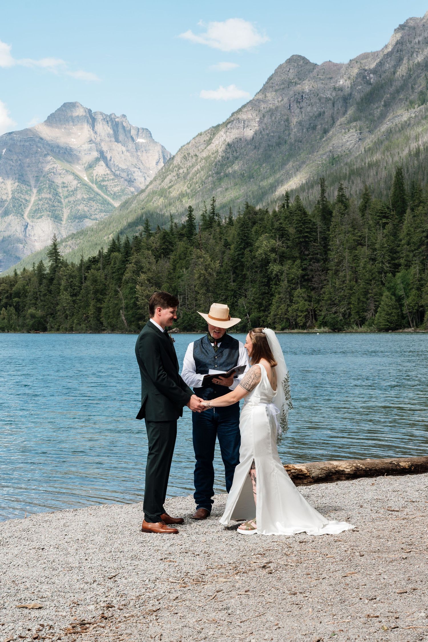 Outdoor wedding ceremony by a mountain lake with bride, groom, and officiant in cowboy hat, surrounded by pine trees.
