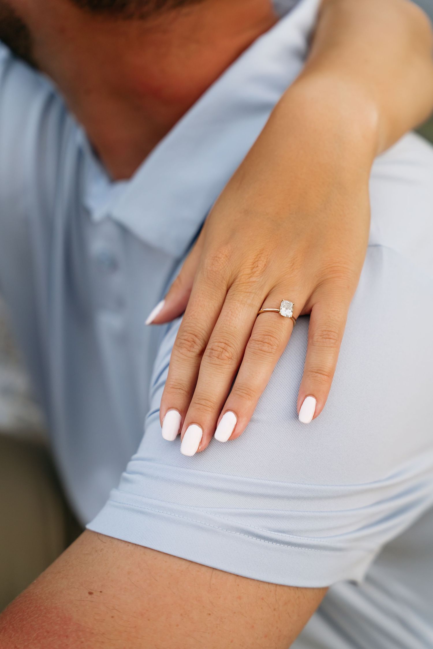 Close-up of hands with white manicured nails and diamond engagement ring on light gray fabric background.
