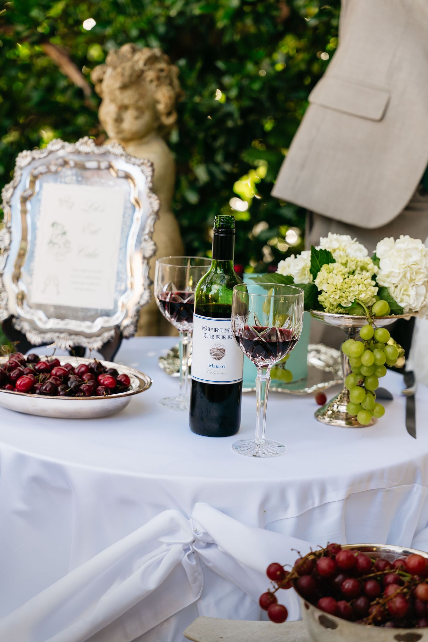 Elegant outdoor table setting with white linens, wine glasses, fresh grapes, and ornate silver serving pieces.