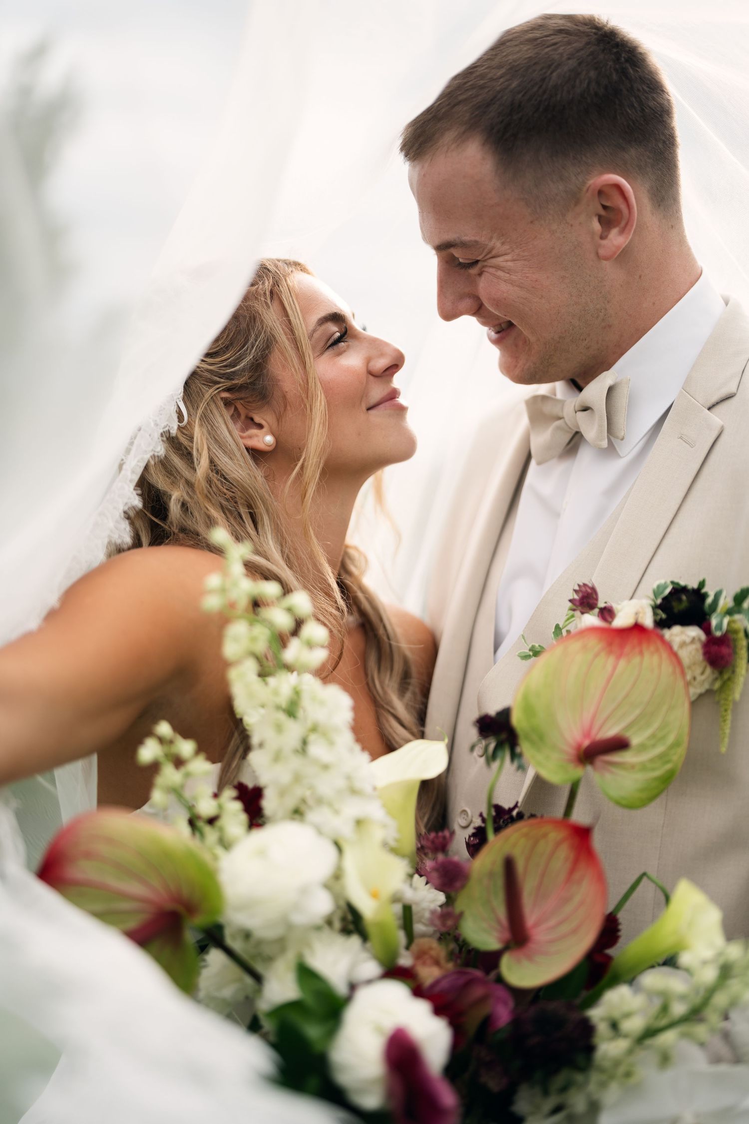 Smiling bride gazes up at groom beneath a veil, with a lush floral bouquet of calla lilies in the foreground.