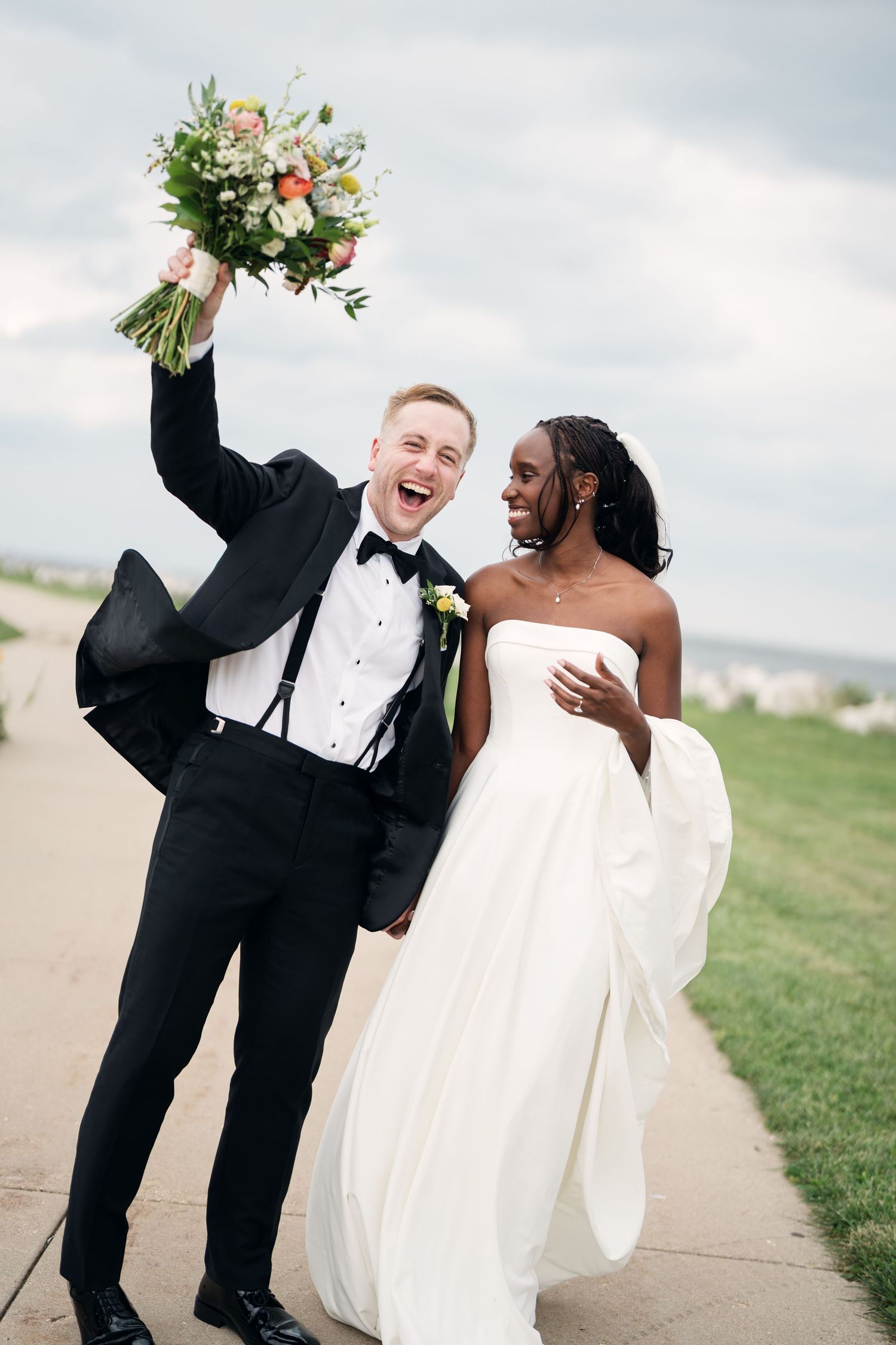 Joyful groom raises bouquet while bride laughs beside him in elegant strapless wedding gown.