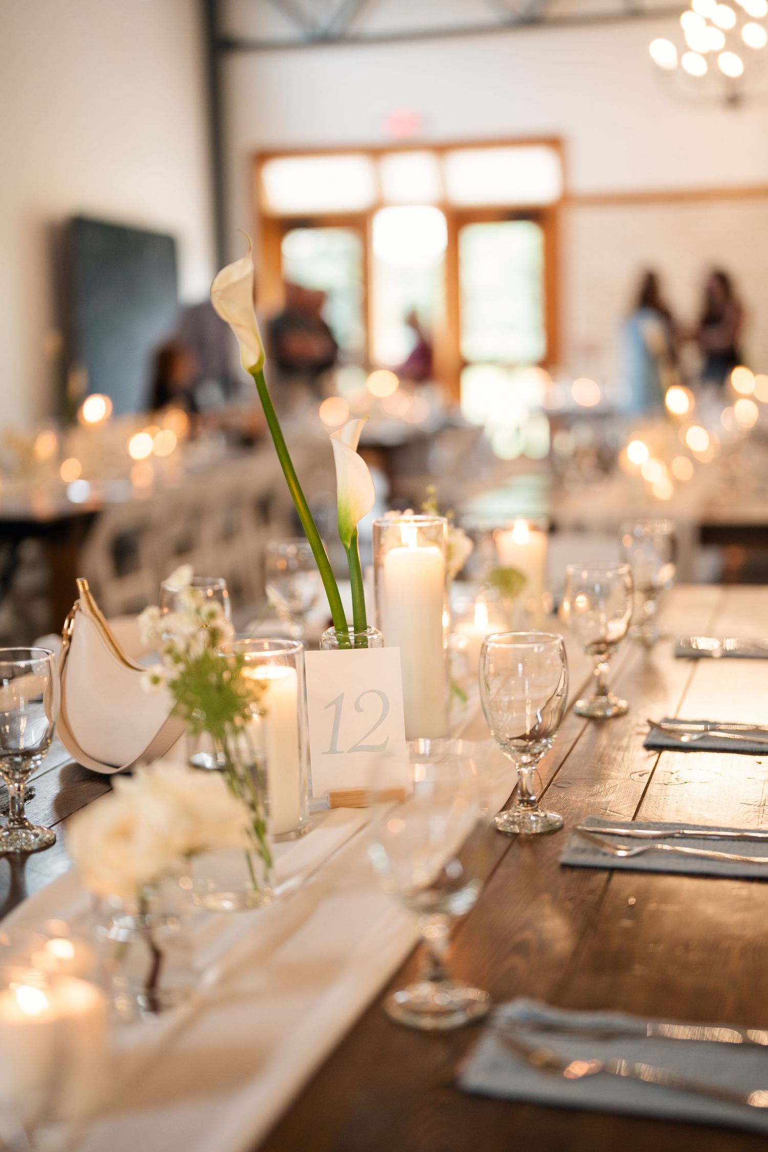 Elegant wedding reception table with calla lily centerpieces, white candles, and glassware on rustic wooden farm table.