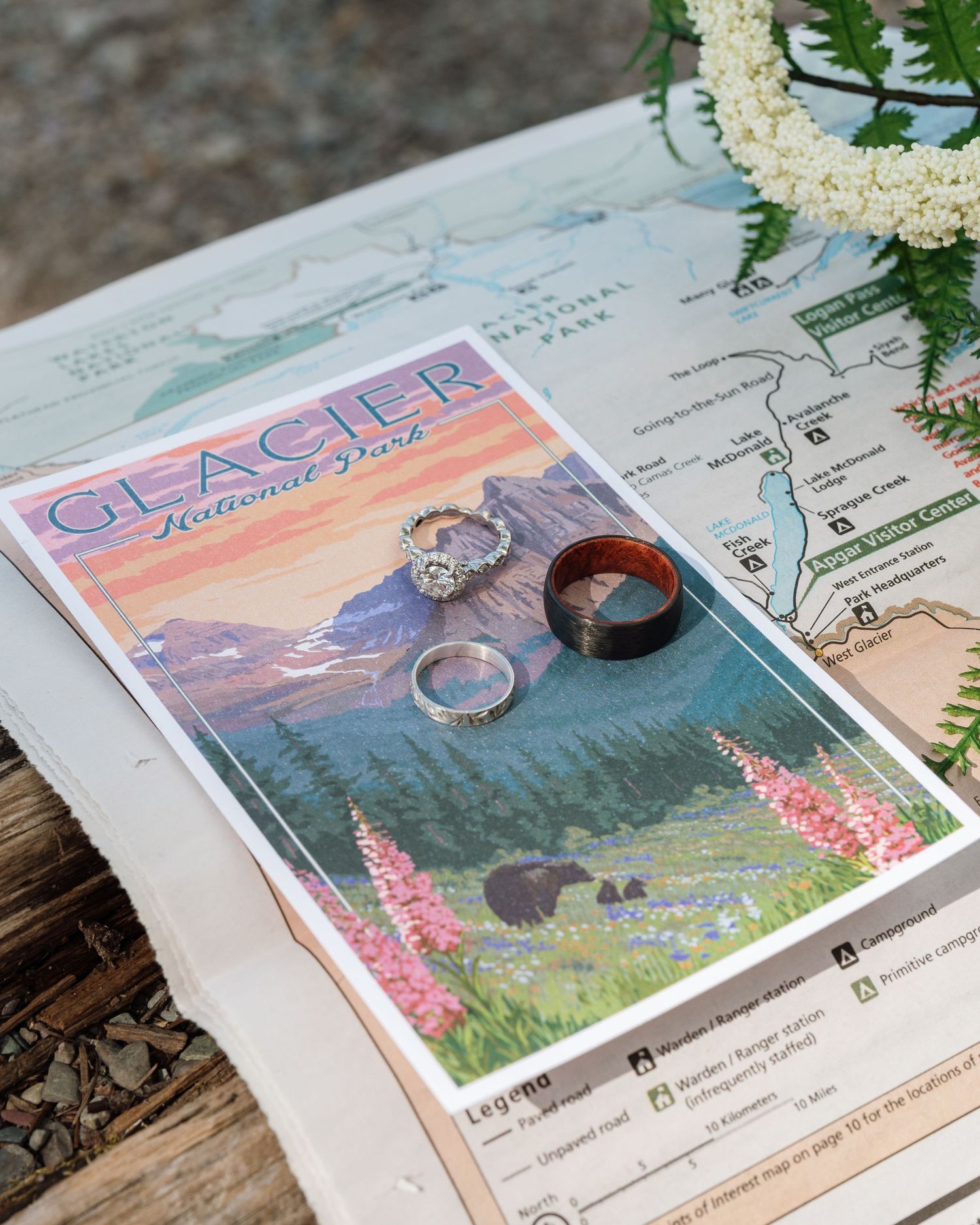 Wedding rings displayed on a Glacier National Park postcard atop a park map, with a floral lei nearby.