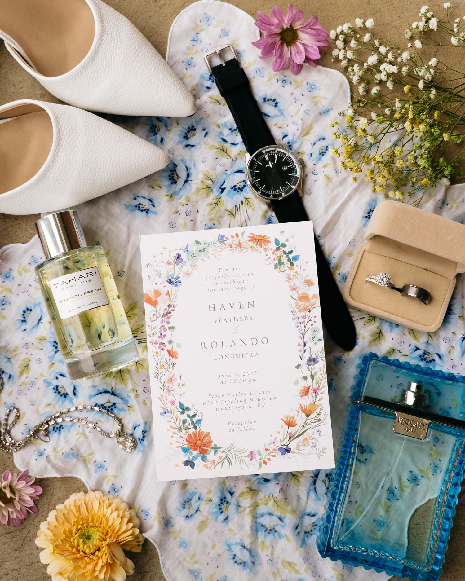 Wedding details flatlay with floral invitation, white heels, perfume, watch, jewelry box and flowers on blue floral fabric.