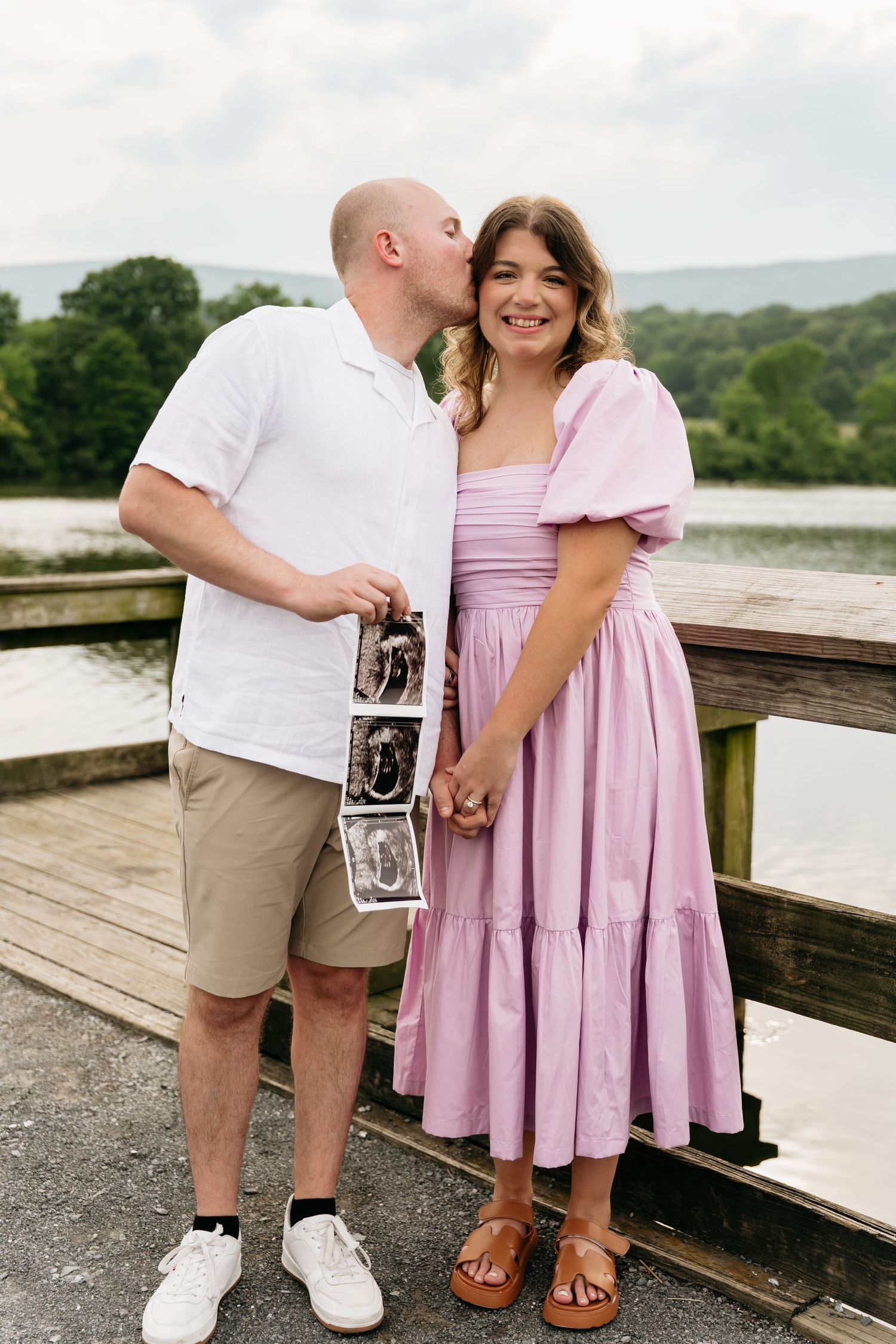 Couple standing on wooden deck holding ultrasound photos in casual summer outfits near water and trees in background.
