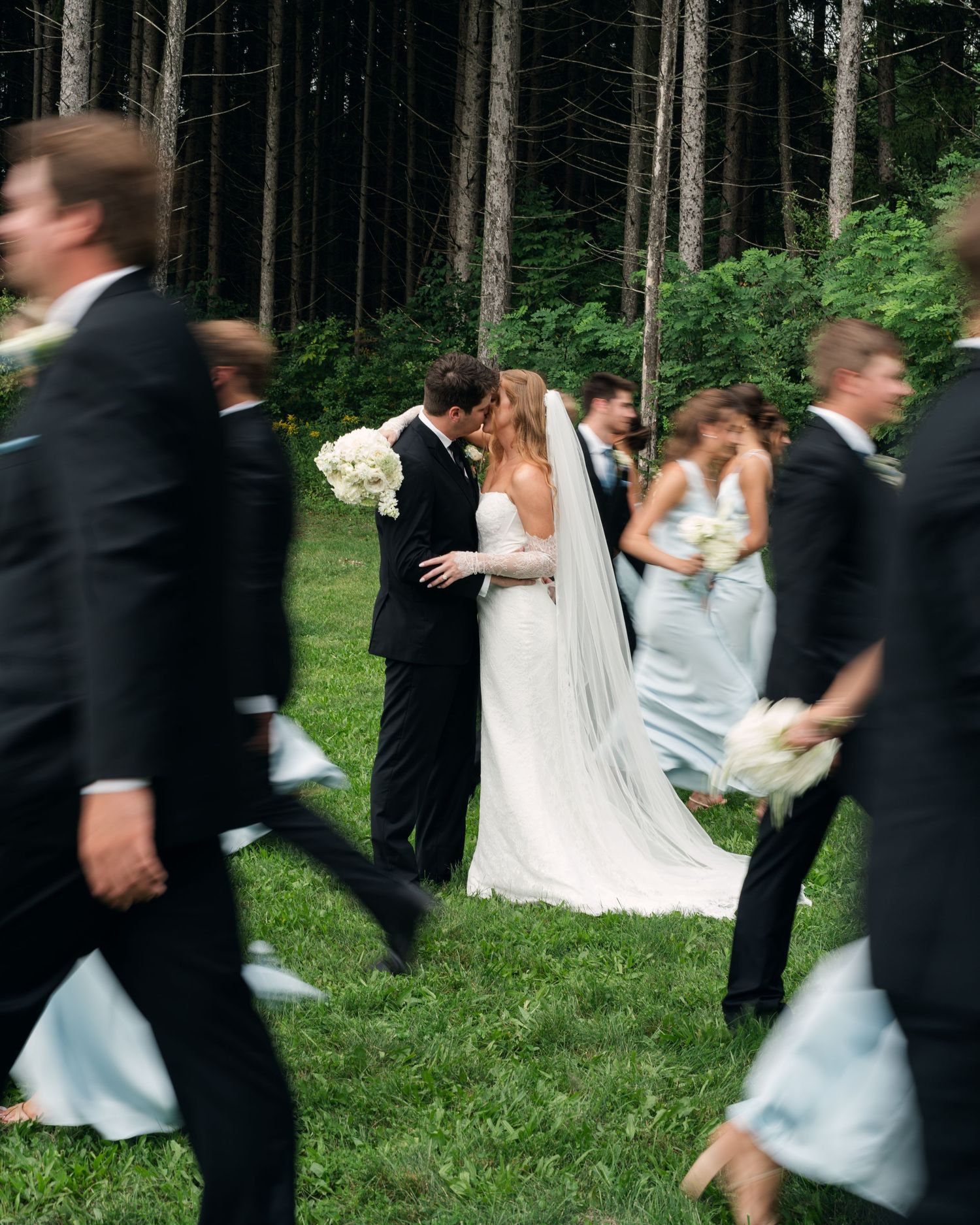 Bride and groom share a kiss during outdoor wedding ceremony while bridal party walks past in black suits and blue dresses.