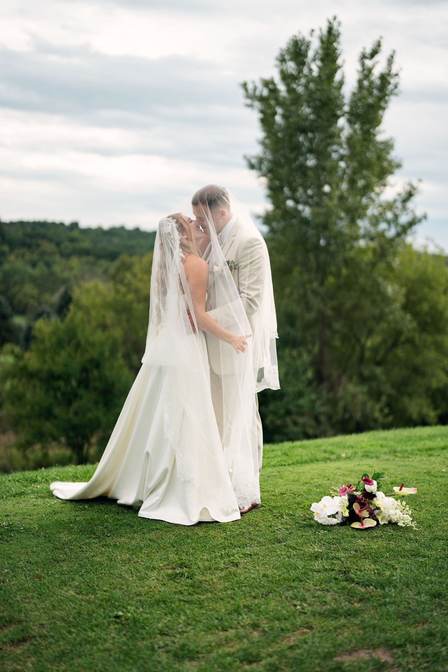 Bride and groom share a romantic kiss under a flowing white veil on a lush green lawn at an outdoor wedding.