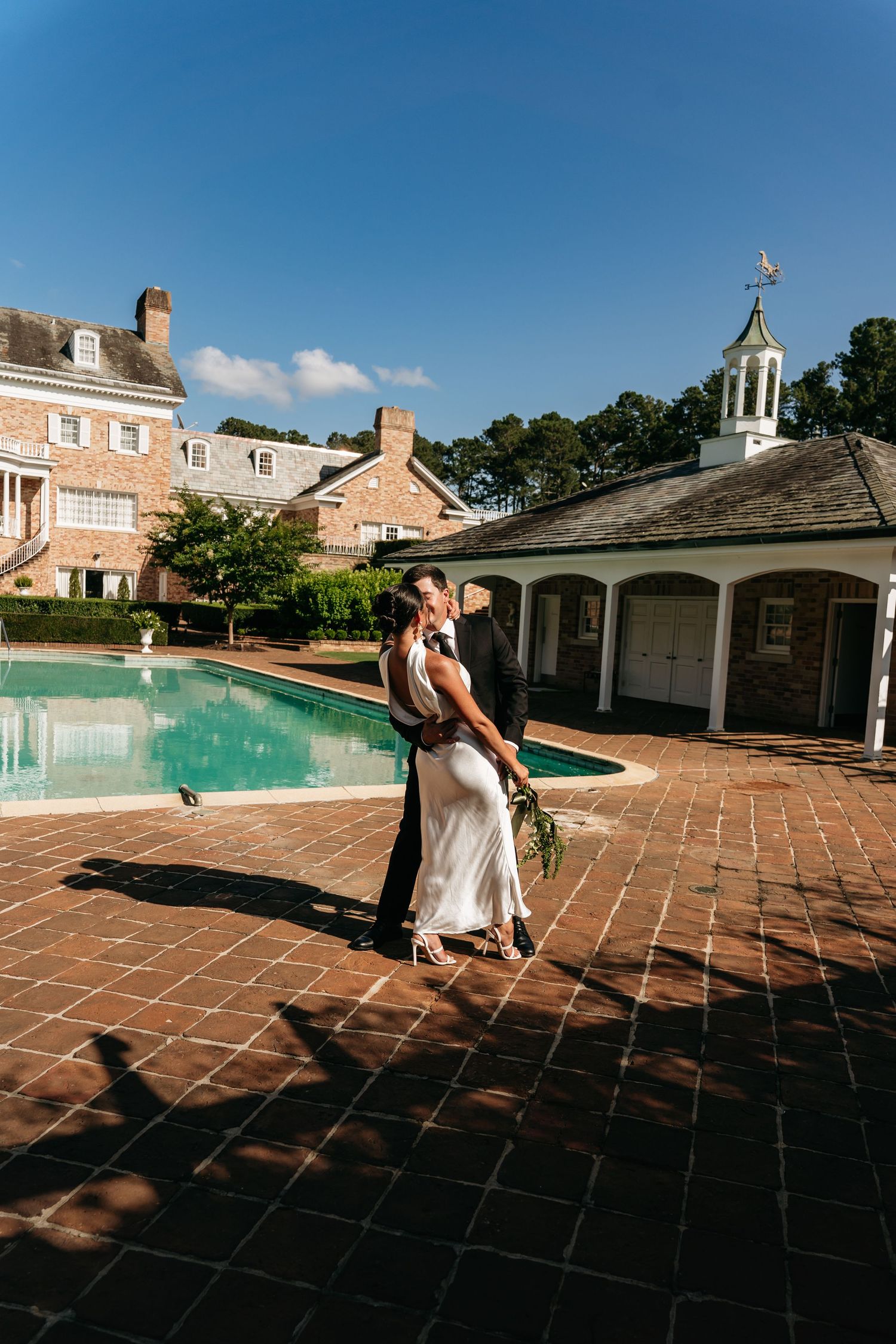 Couple dancing and twirling on a terracotta tile patio near a swimming pool at an elegant estate on a sunny day.