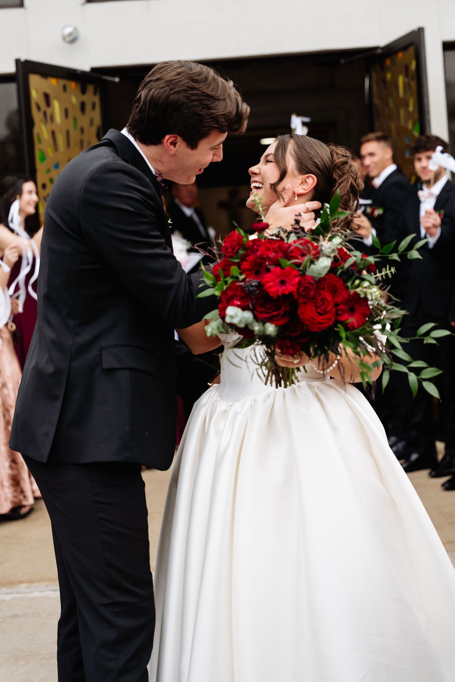 Joyful bride in white ball gown holding red roses bouquet laughs with groom in black tuxedo at wedding reception.
