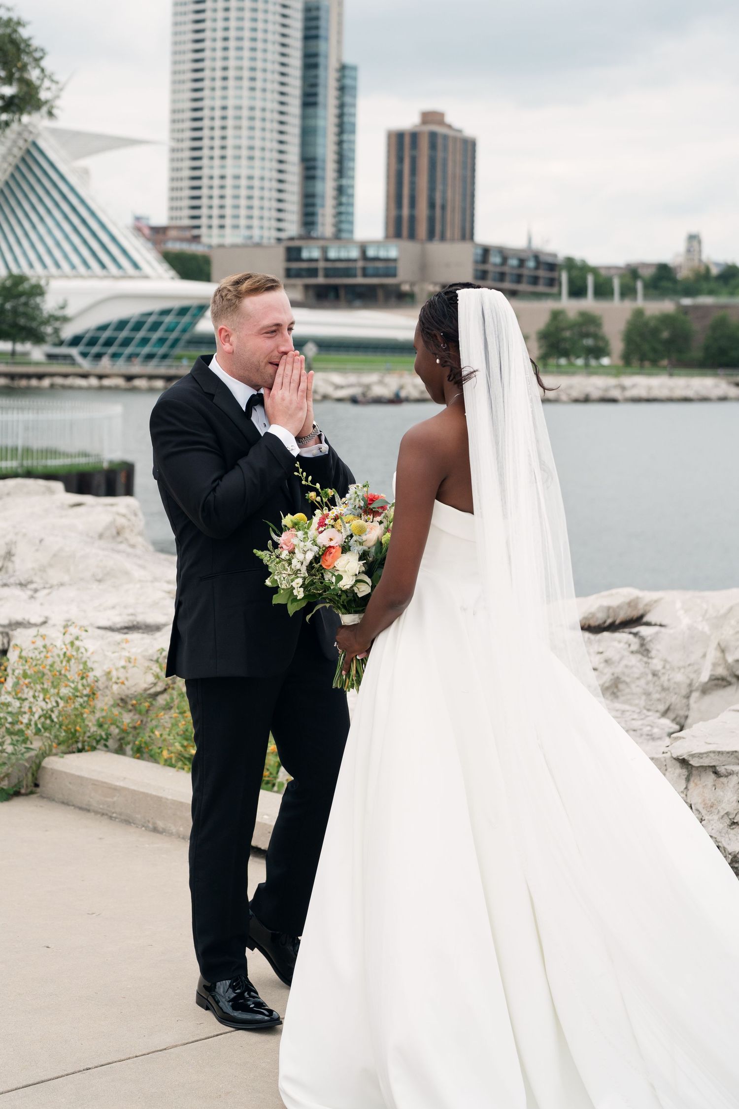Emotional groom reacts with clasped hands as bride in white gown holds bouquet during first look by waterfront.